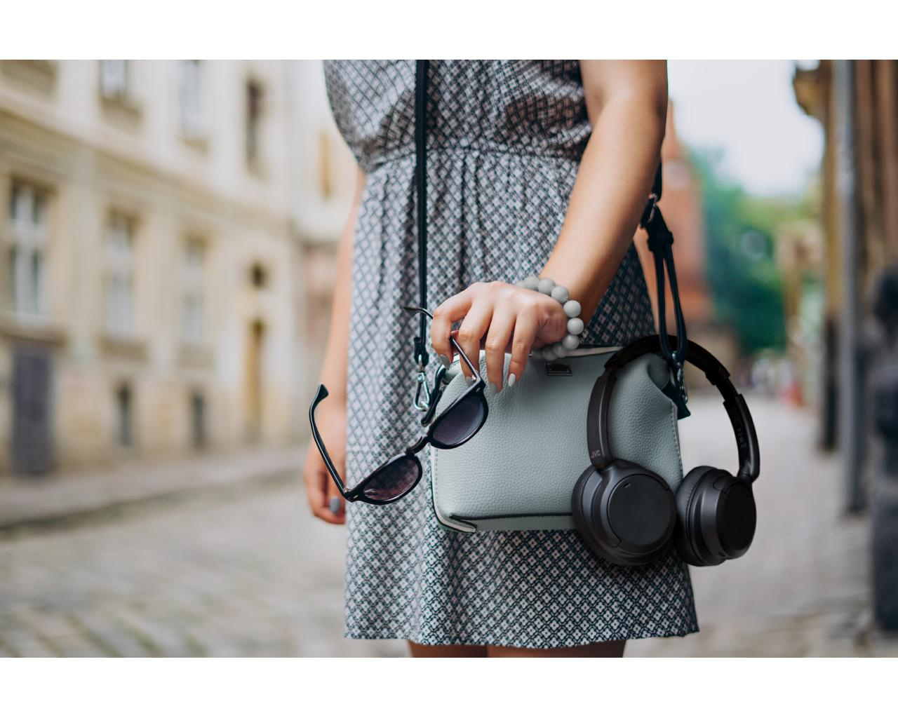 Kobieta trzymająca torebkę ze słuchawkami i okularami przeciwsłonecznymi na ulicy w słońcu.