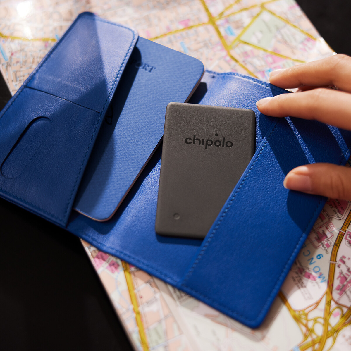 A hand opening a blue wallet, next to a passport and a gray Chipolo tracker.