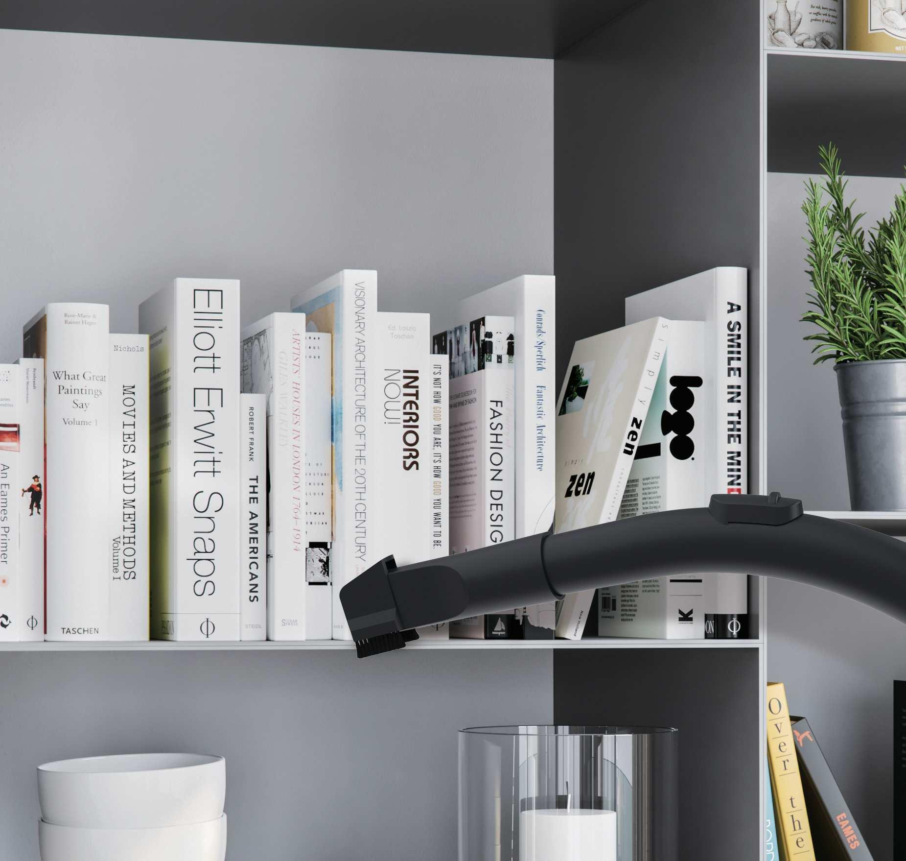 Książki na półce czyszczone odkurzaczem. Scena jest w pomieszczeniu i oświetlona naturalnym światłem.