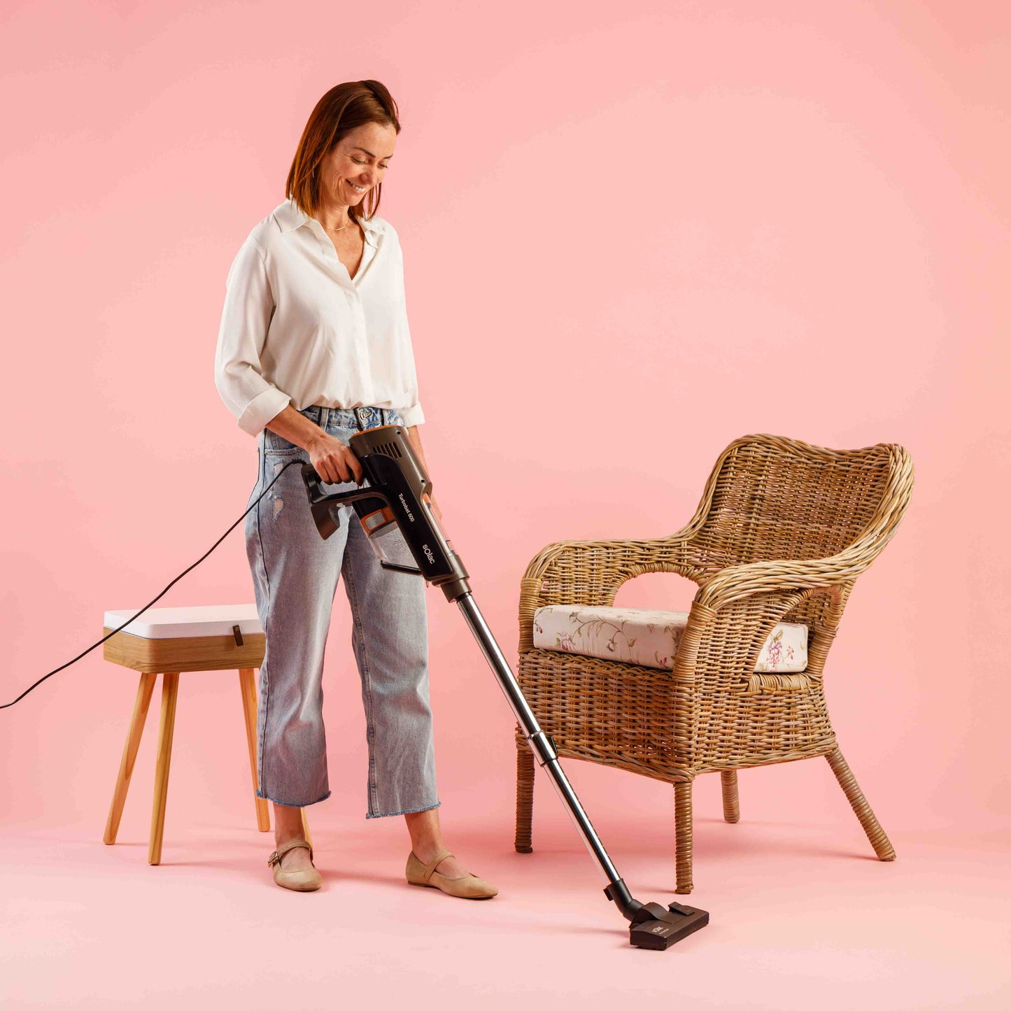 Kobieta odkurza pokój z różowym tłem. Obecny jest fotel i stolik.