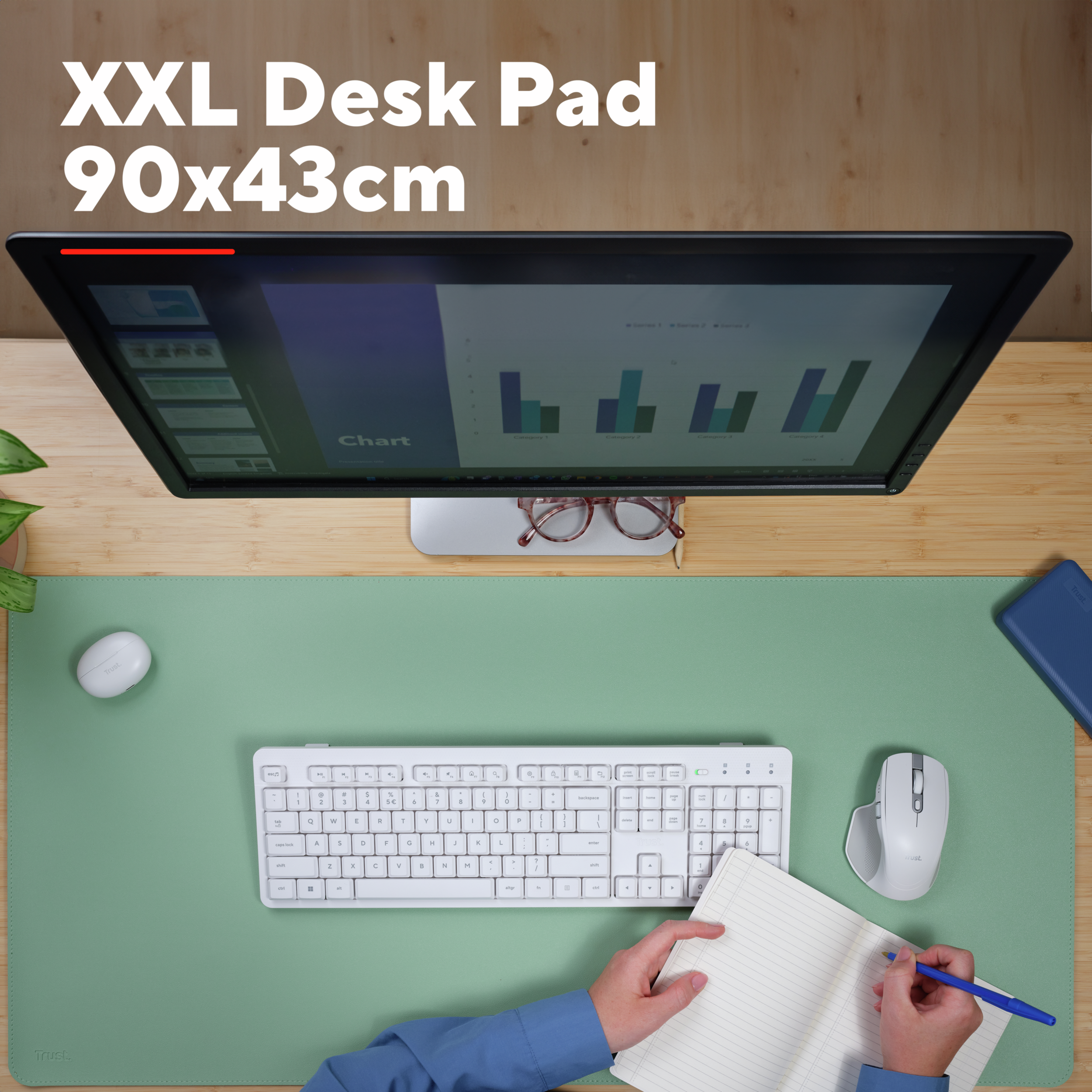 A computer setup with desk pad, monitor, keyboard, mouse, glasses, and a notepad. A person is writing.