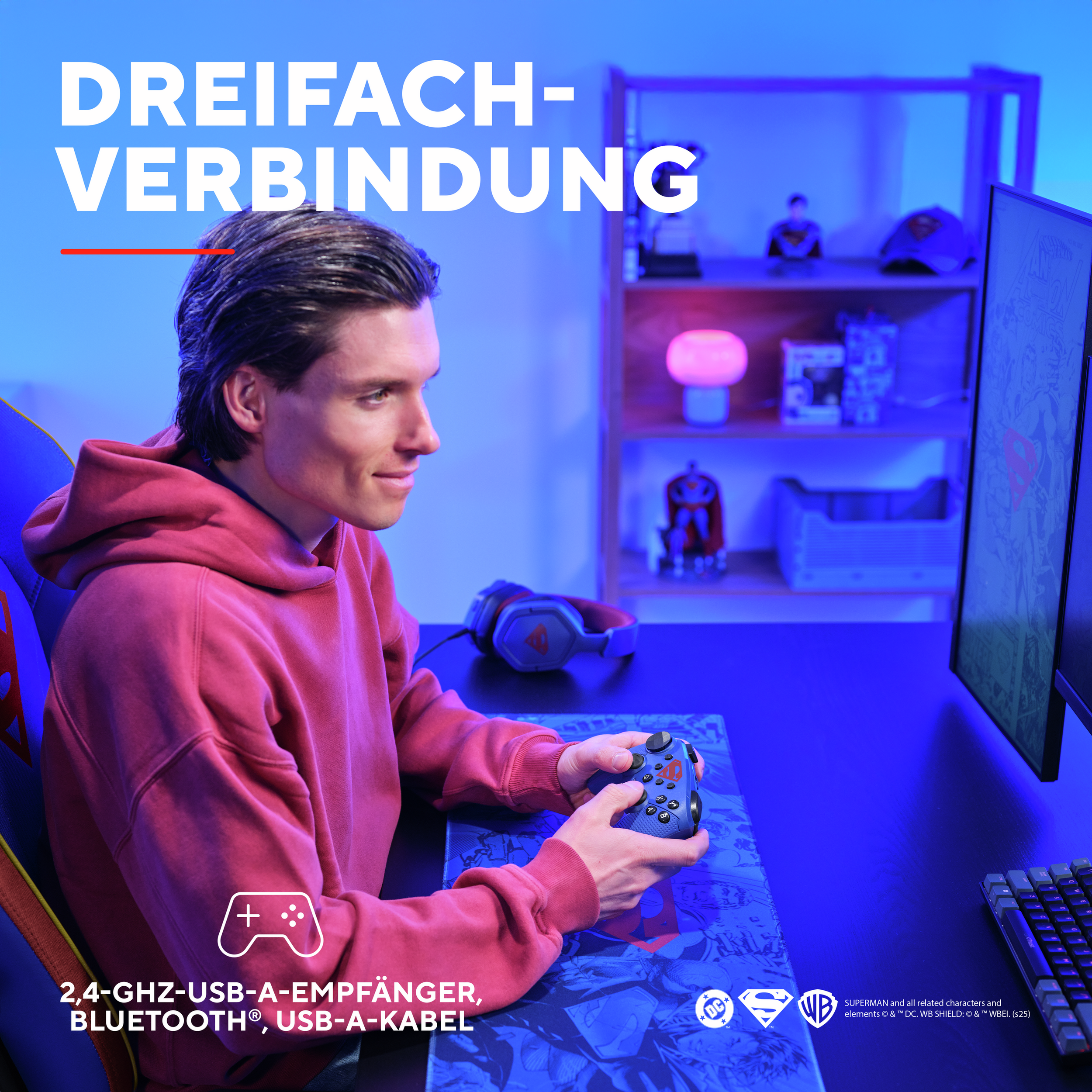 Un uomo tiene in mano un controller di gioco blu con il logo di Superman. Dietro ci sono una scrivania, un monitor e delle figure.
