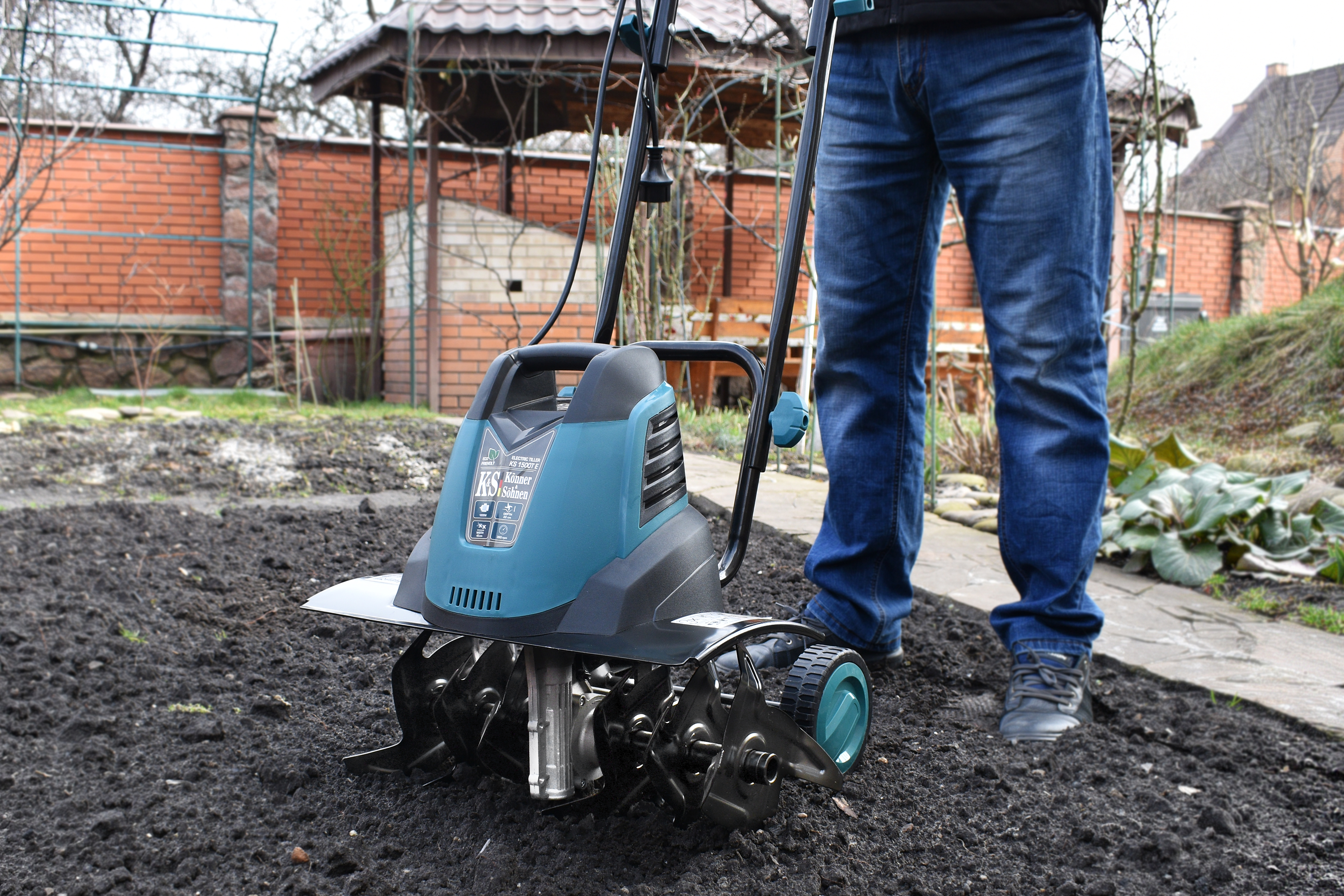 Persona che utilizza una motozappa turchese e nera in un giardino. Il terreno è scuro, con sfondo in mattoni.