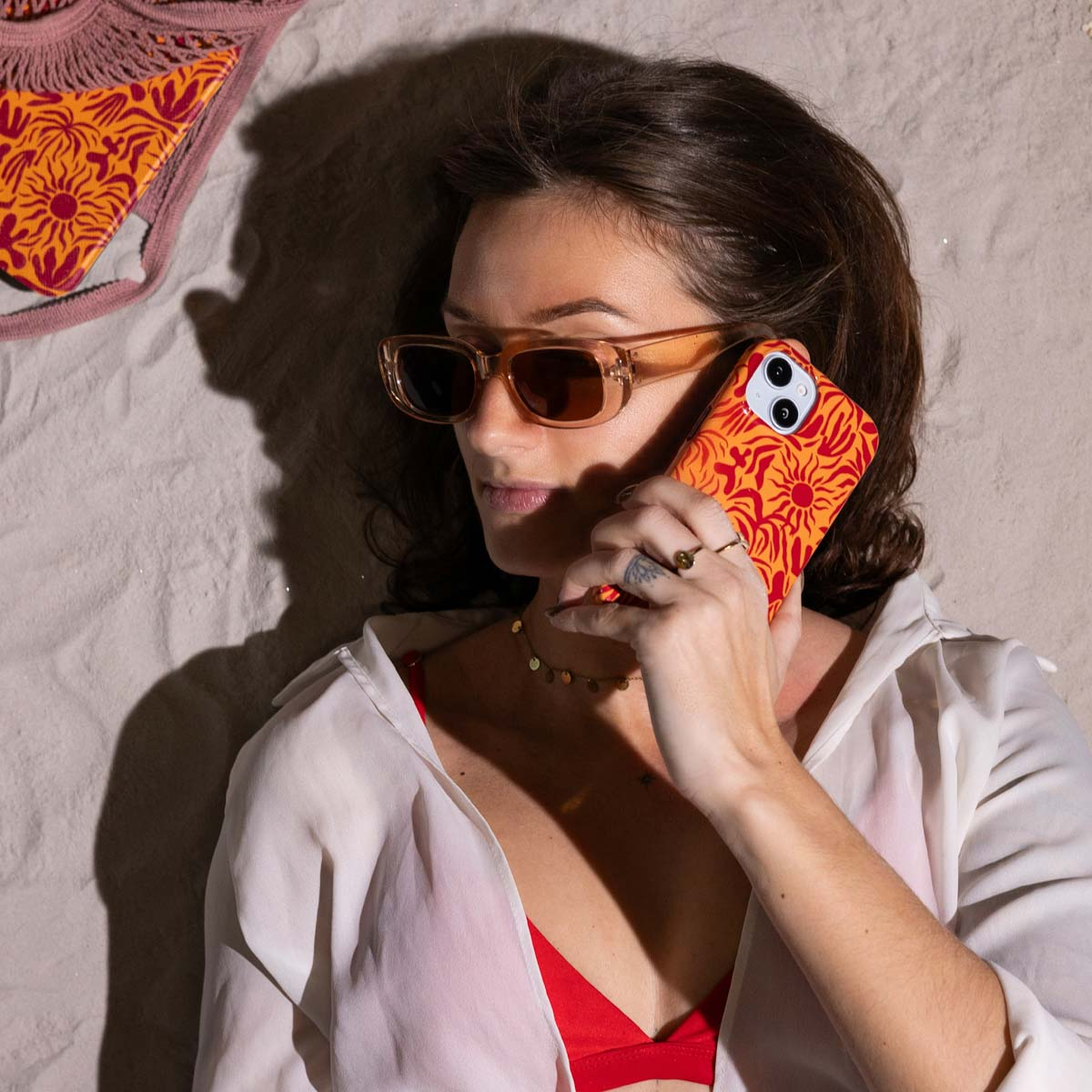 Femme sur le sable portant des lunettes de soleil, tenant un téléphone avec une coque florale orange. Bikini et sac présents.