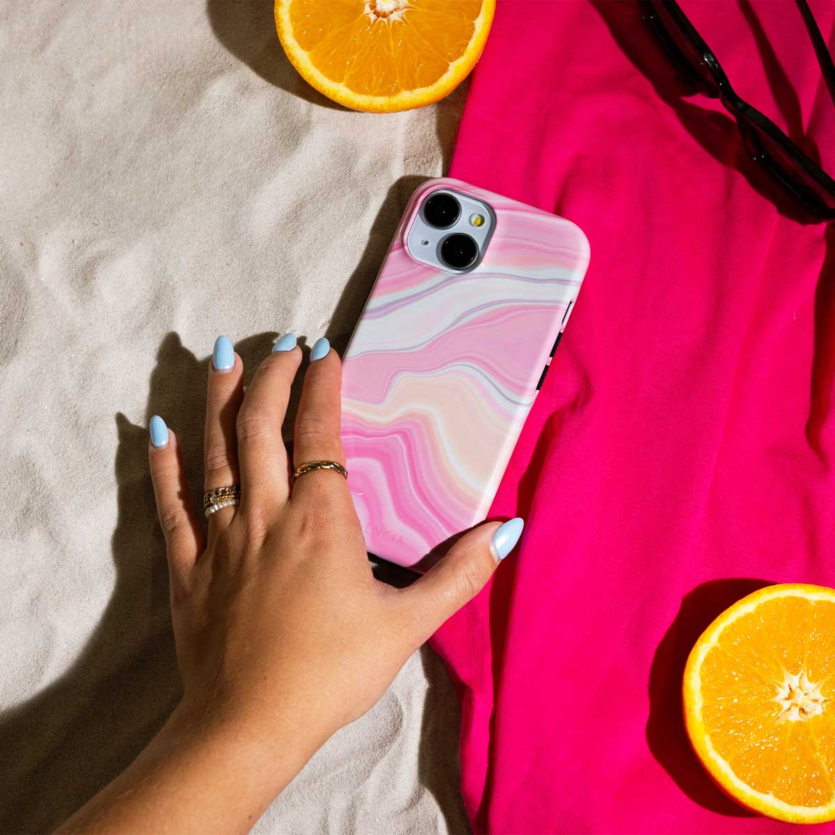 Hand die een roze en witte marmeren telefoonhoes vasthoudt. Sinaasappelschijf en zonnebril aan de zijkant.