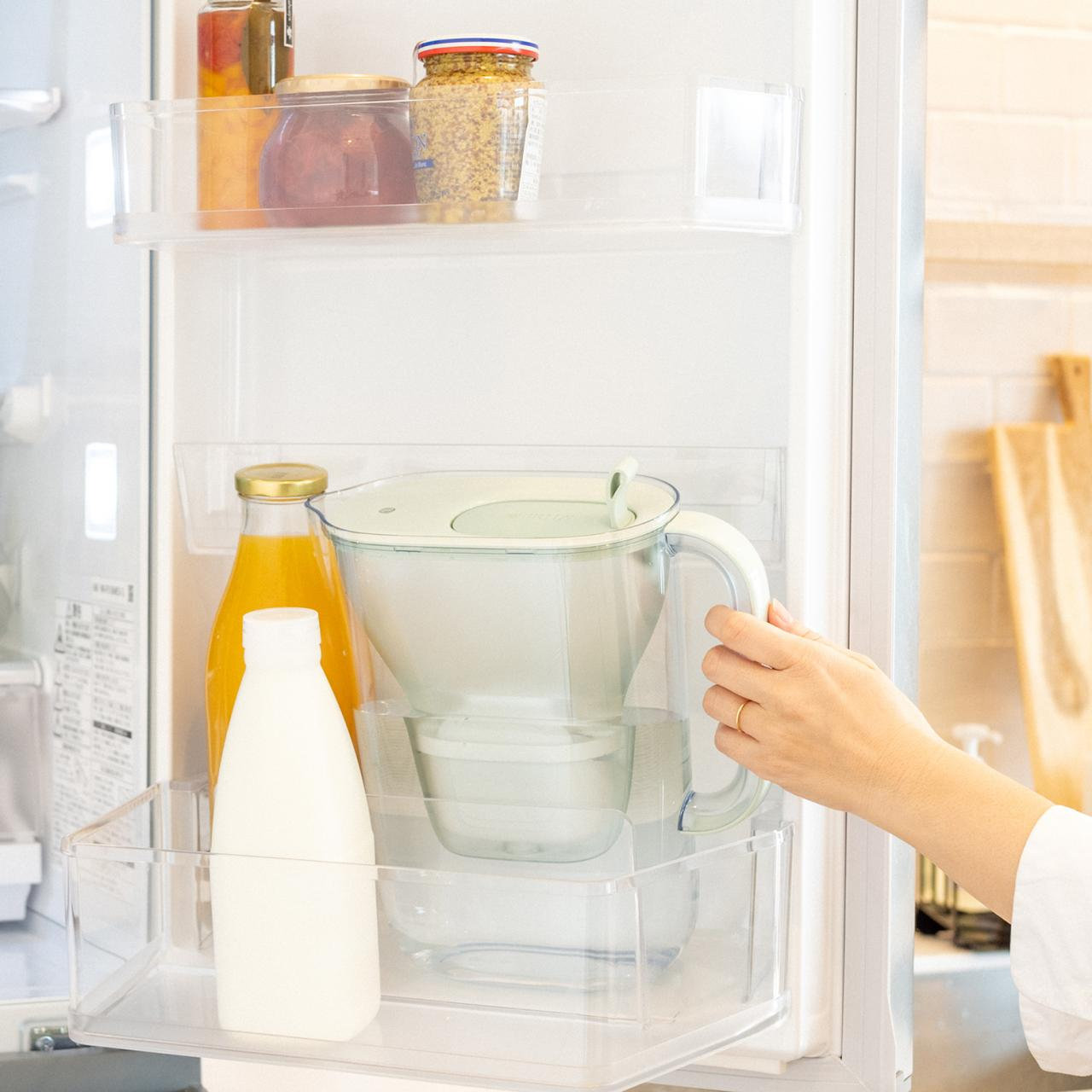 Wnętrze lodówki z sokiem, mlekiem, dzbankiem z filtrem wody i sięgającą ręką.