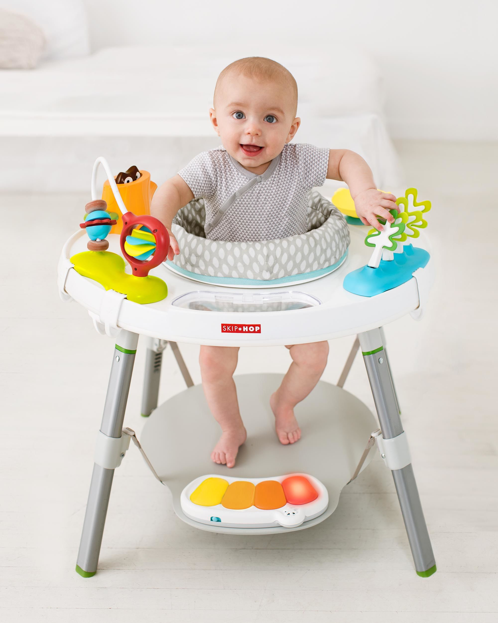Ein Baby sitzt in einem weißen Spieltisch. Spielzeug liegt auf dem Tisch.