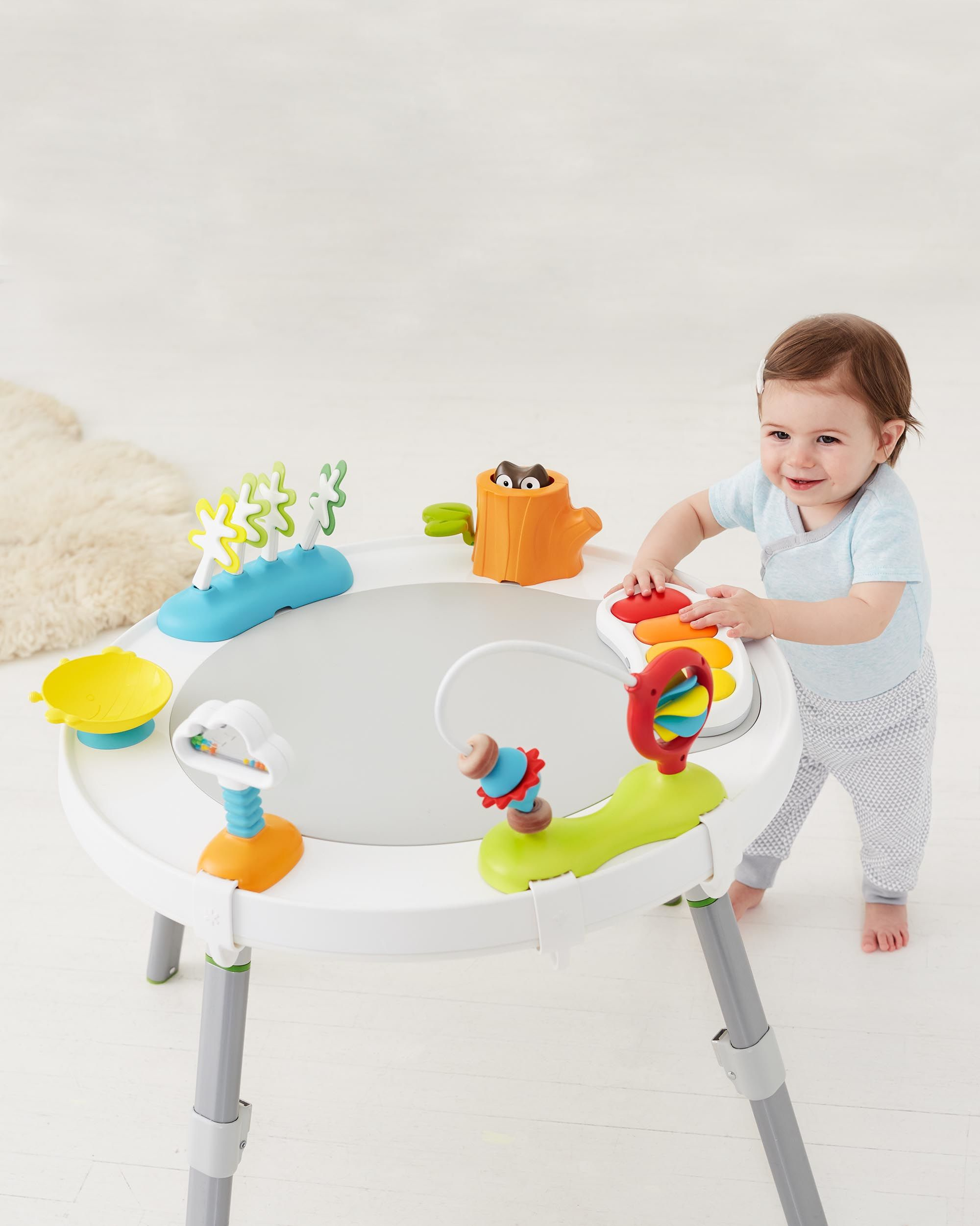 Baby steht an einem Aktivitätstisch mit Spielzeug. Lächelt.