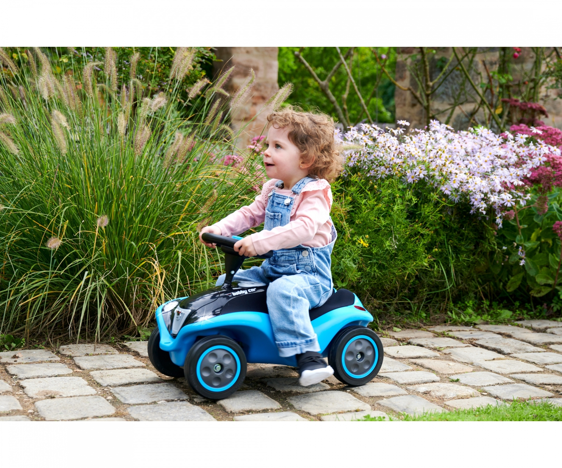 Mała dziewczynka na niebieskim samochodziku, uśmiechnięta. Ma na sobie dżinsowe ogrodniczki i różową koszulę.