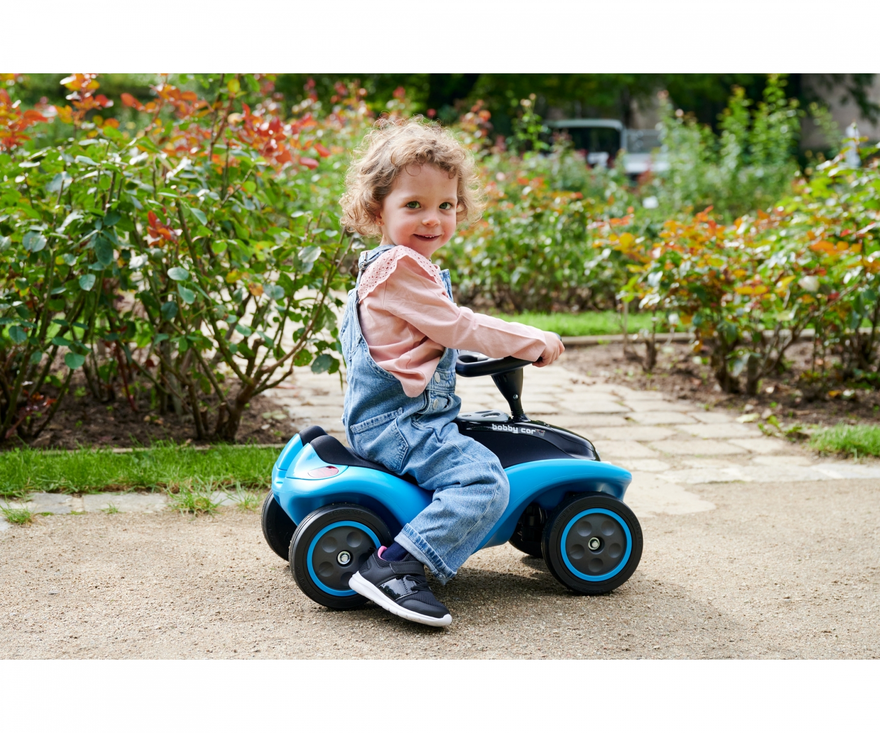 Dziewczynka jedzie niebieskim samochodzikiem po ścieżce. Rośliny i kwiaty w tle.