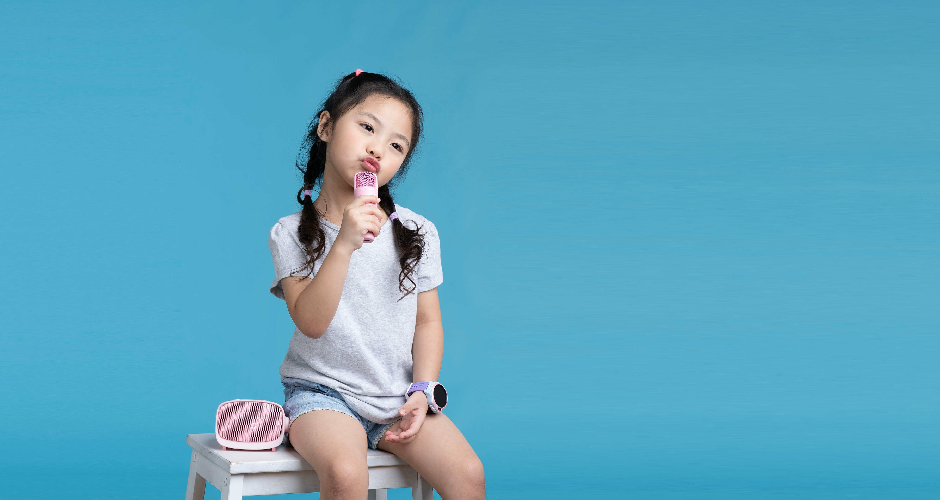 Une fille tenant un microphone rose, assise sur un tabouret blanc devant un fond bleu.