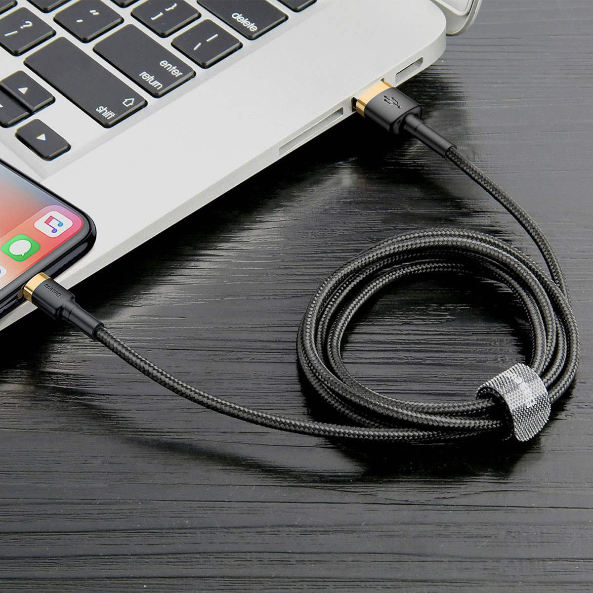 A black cable connected to a phone and a laptop on a wooden surface, with a gray velcro strap.