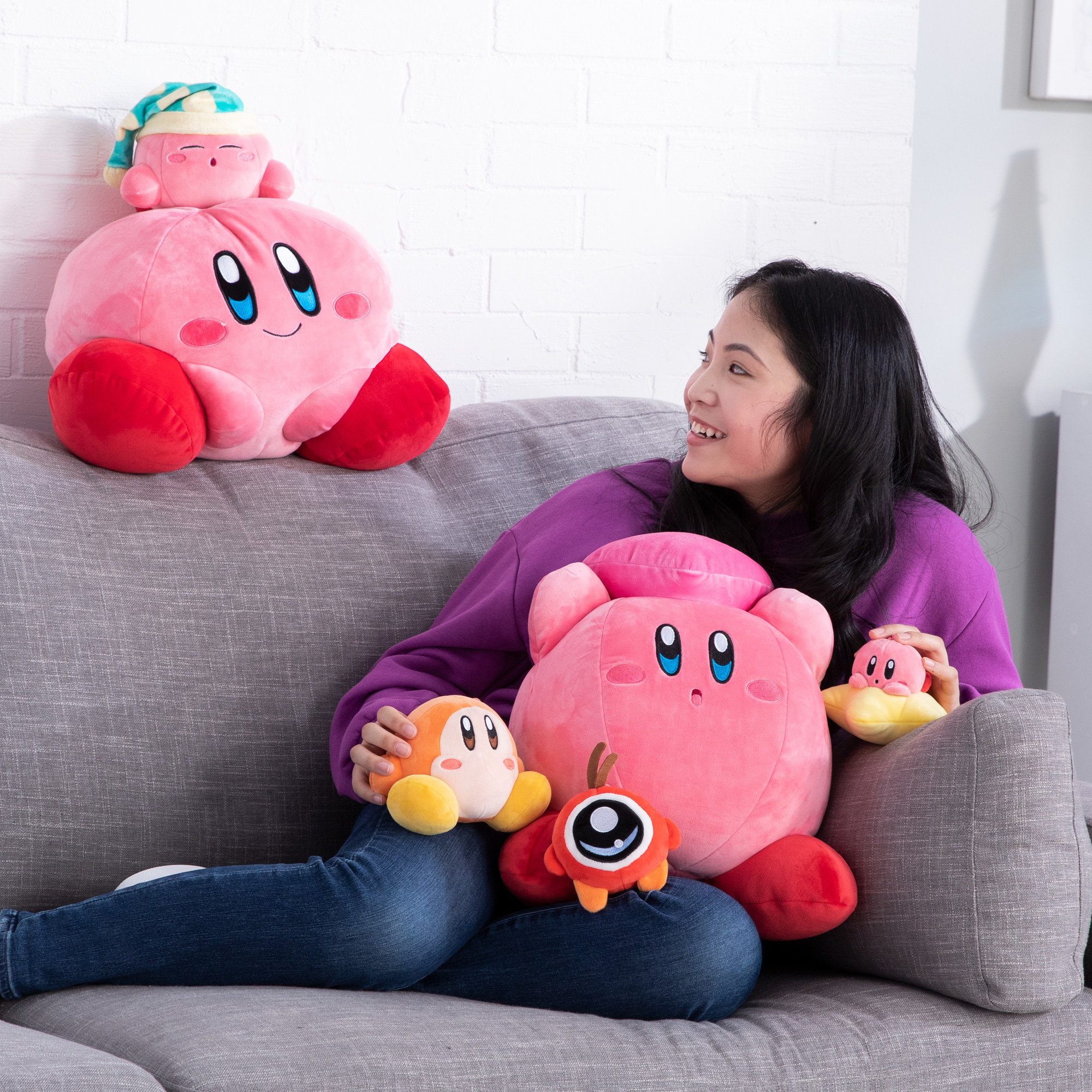 Frau auf einem Sofa, umgeben von Kirby-Plüschtieren, darunter ein großes rosa Exemplar.