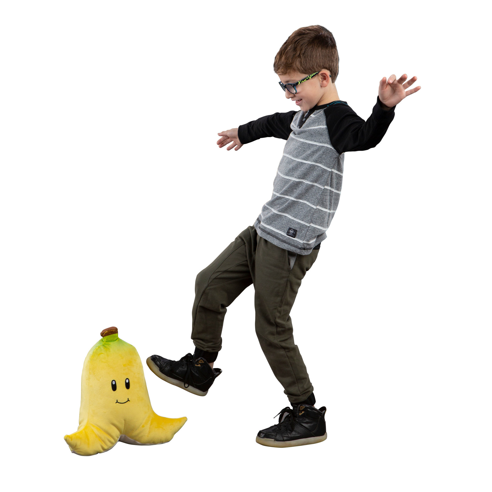 Un garçon donne un coup de pied à une peluche banane souriante. Il porte des lunettes, un t-shirt gris rayé et des chaussures noires.