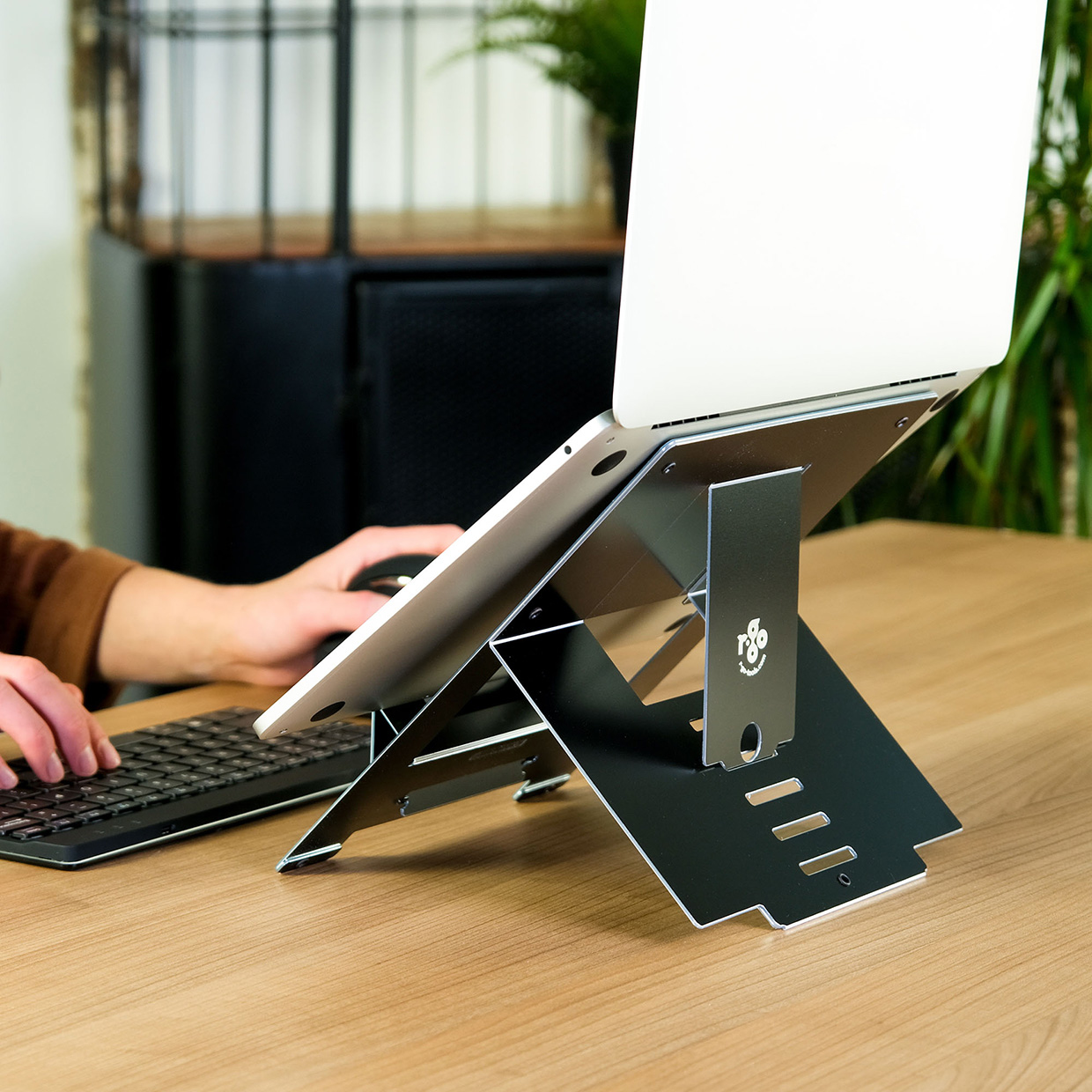 Eine Person benutzt einen Laptop auf einem Ständer. Eine Tastatur und Maus befinden sich auf dem Tisch. Der Ständer ist schwarz.