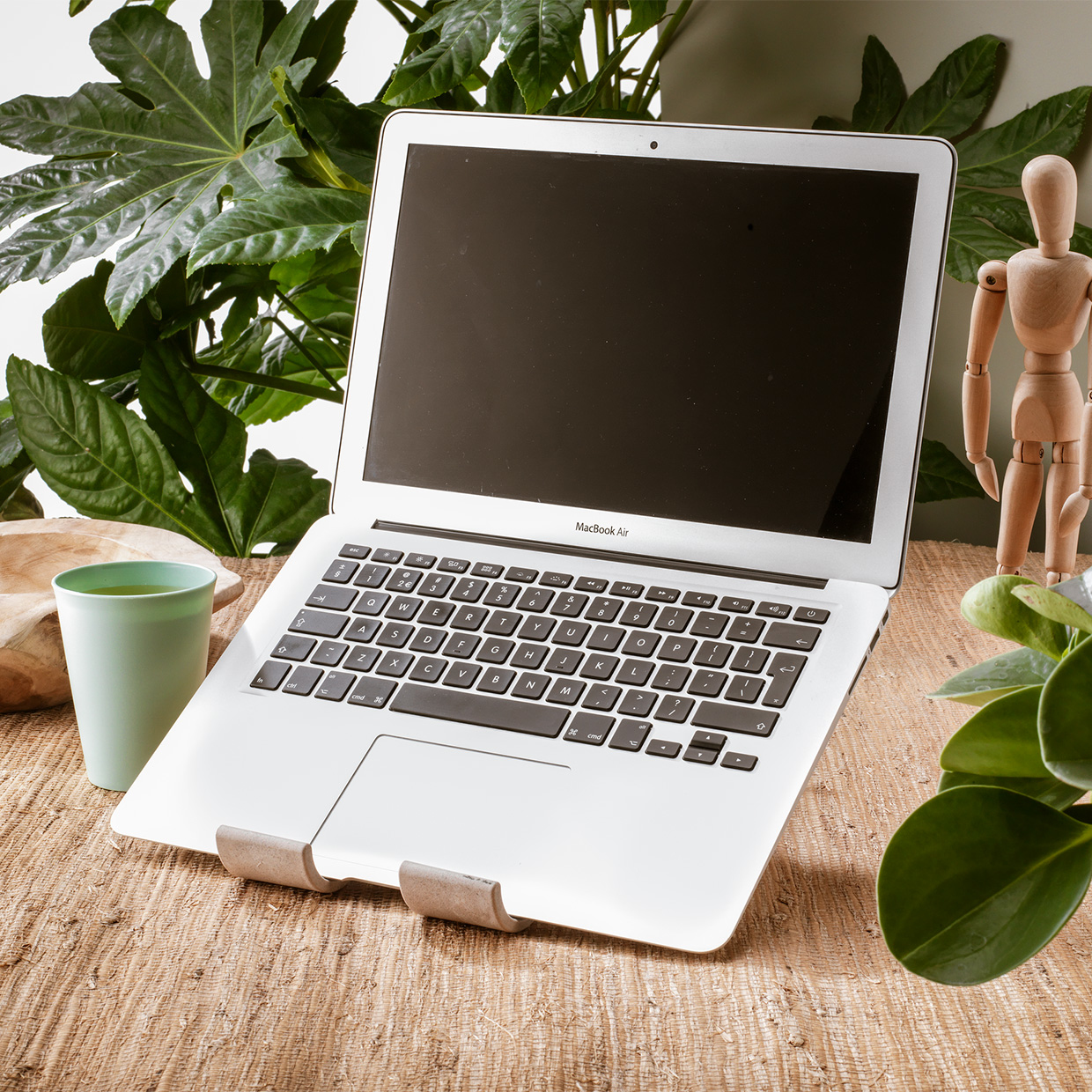 Laptop with black screen on a wooden table, with a green cup and a wooden figure nearby.
