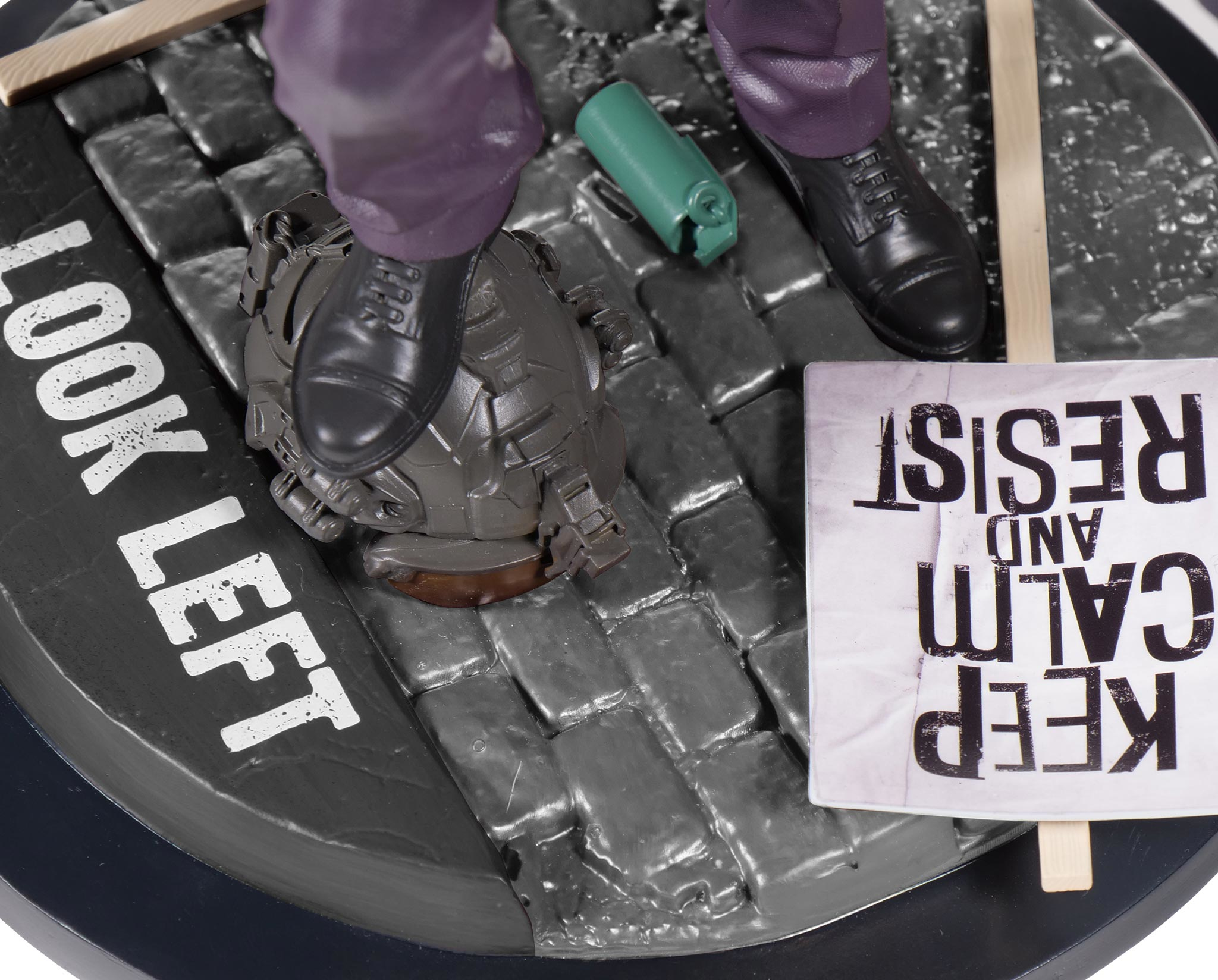 Primer plano de los pies de una estatua en adoquines. Cartel dice 'KEEP CALM AND RESIST'. Lata verde y cartel.