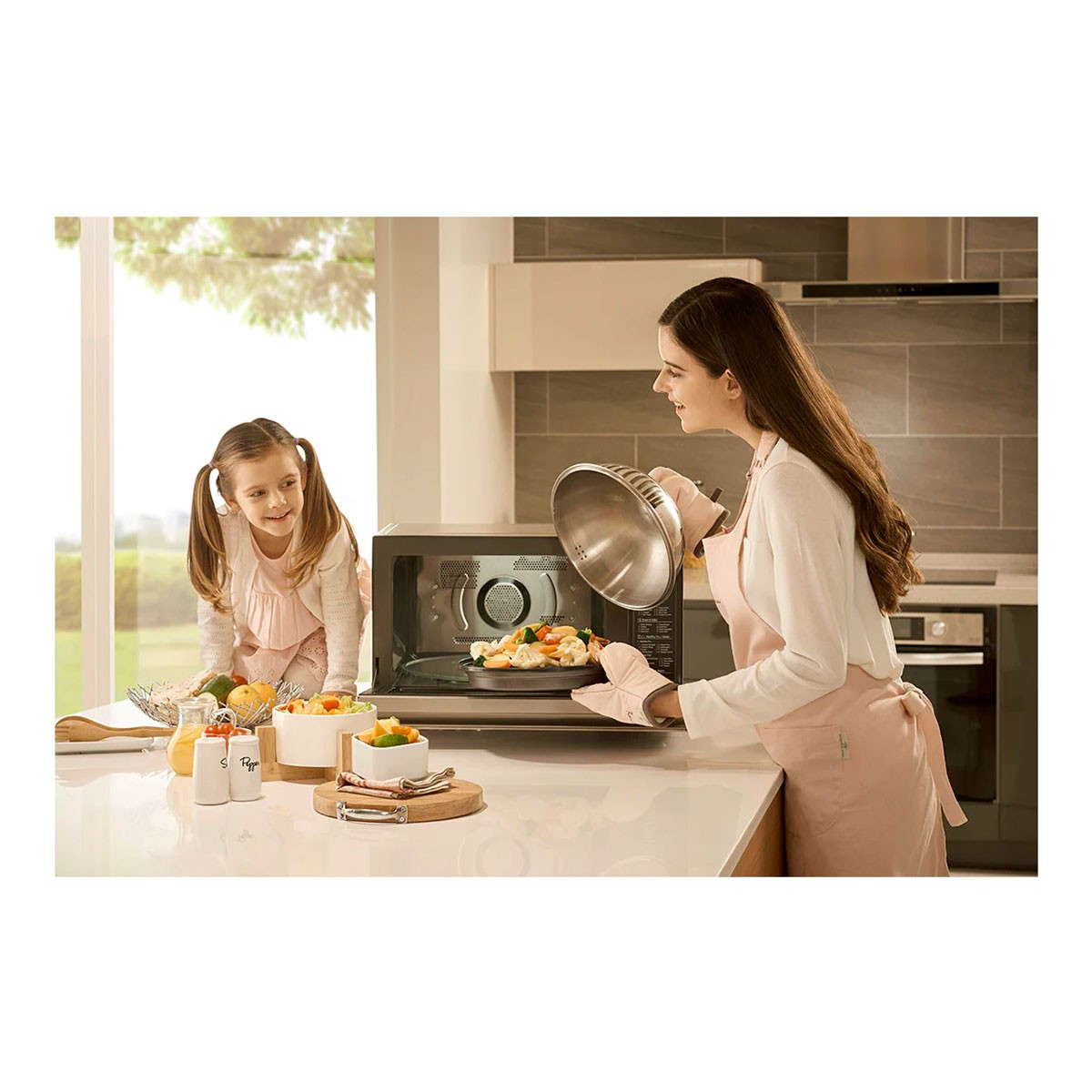 Kobieta wyjmuje jedzenie z kuchenki mikrofalowej. Dziewczynka patrzy. Warzywa i owoce na blacie. Otoczenie kuchni.