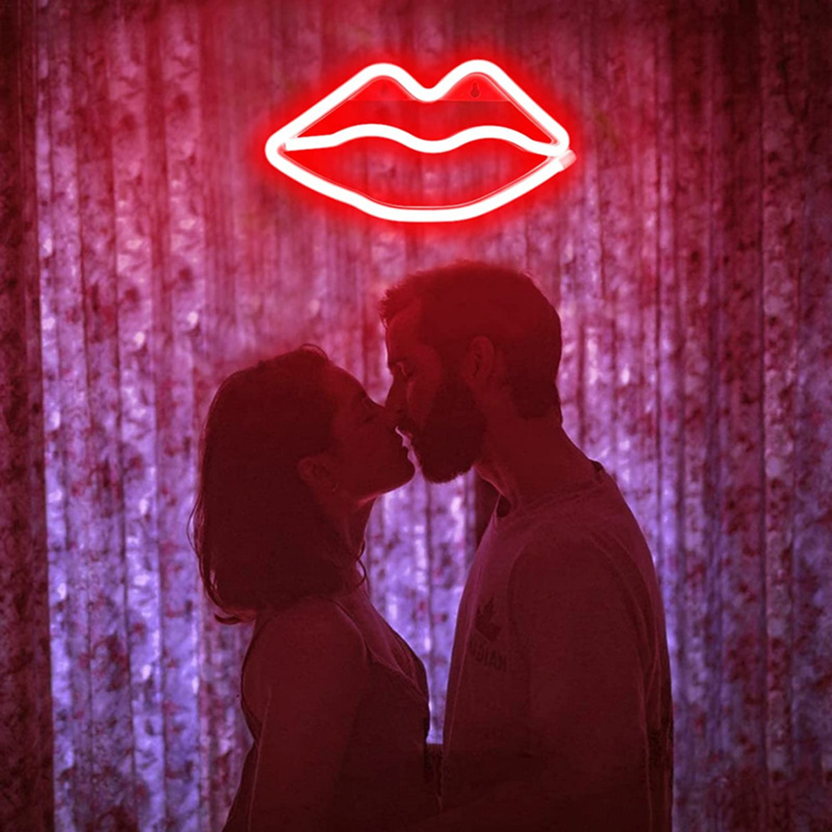 Coppia che si bacia davanti a un'insegna di labbra al neon rosse. Lo sfondo è una tenda rosso scuro sfocata.