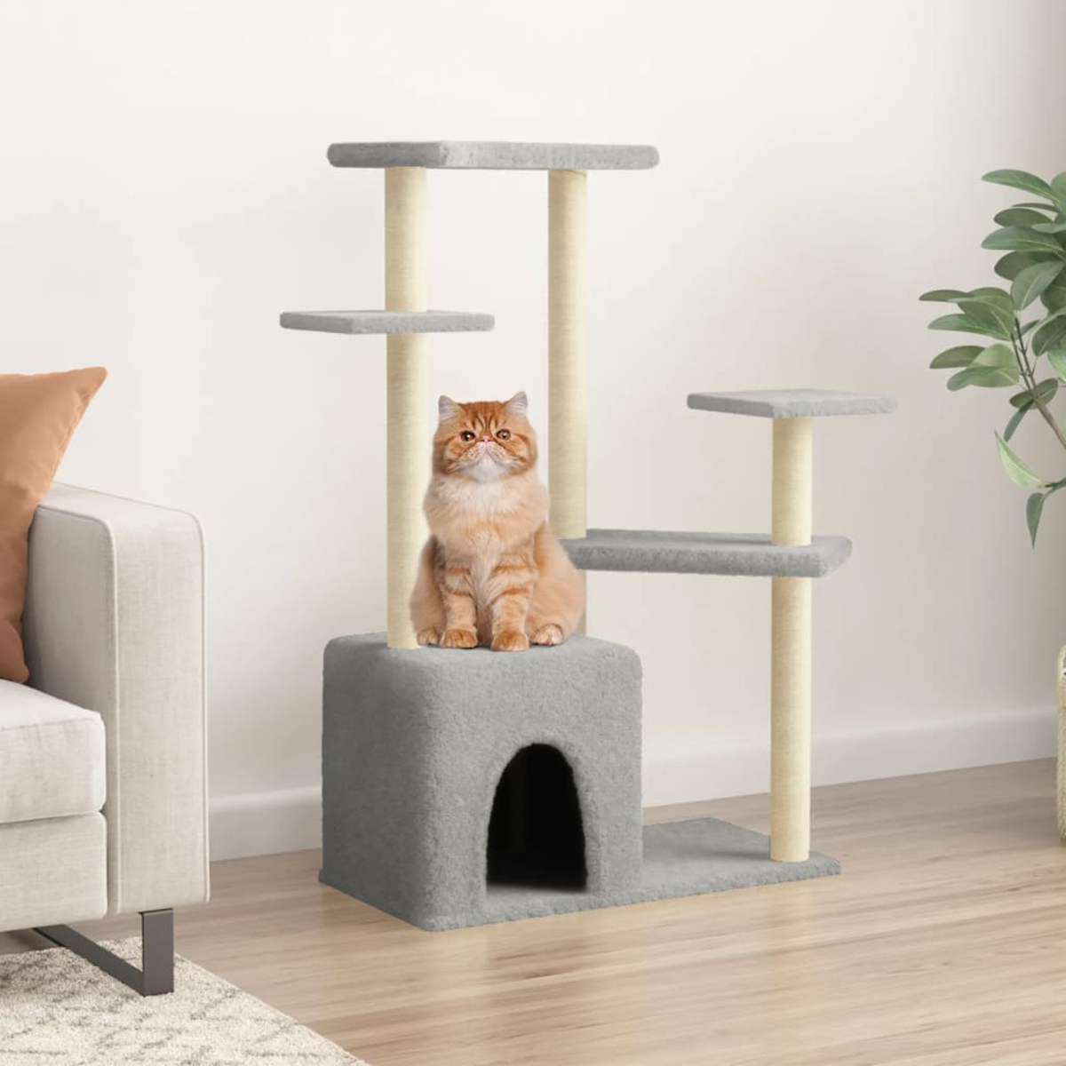 Eine orange Katze sitzt auf einem grauen Kratzbaum in einem Wohnzimmer. Beige Sofa im Hintergrund.