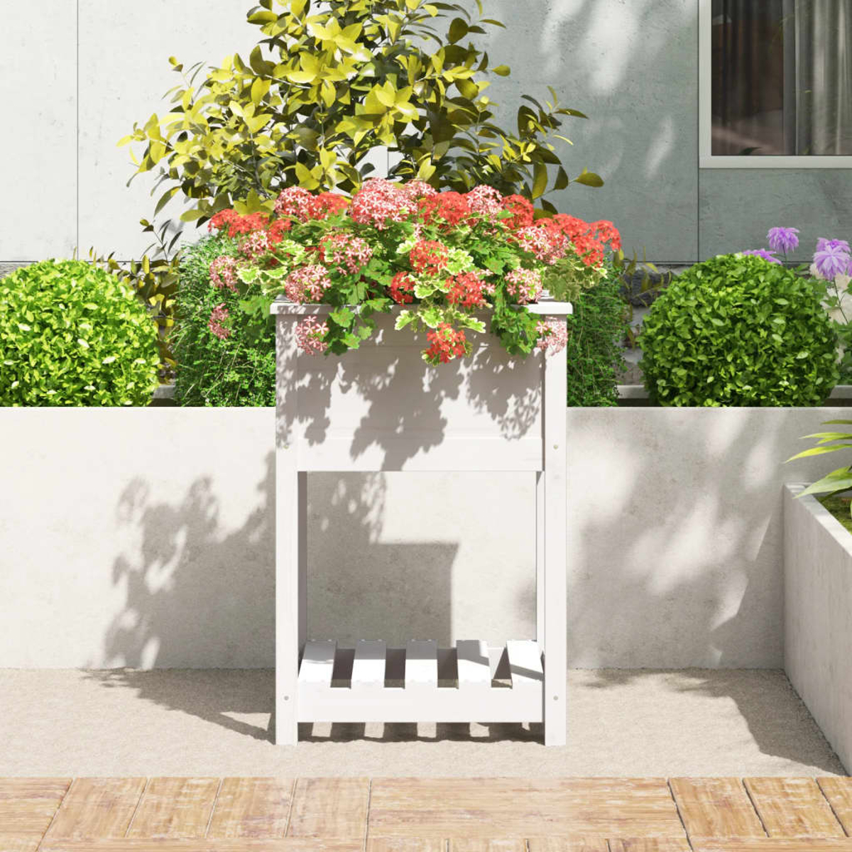 Eine weiße Pflanzkasten mit roten und rosa Blumen. Auf einer Terrasse mit grünen Büschen im Hintergrund.