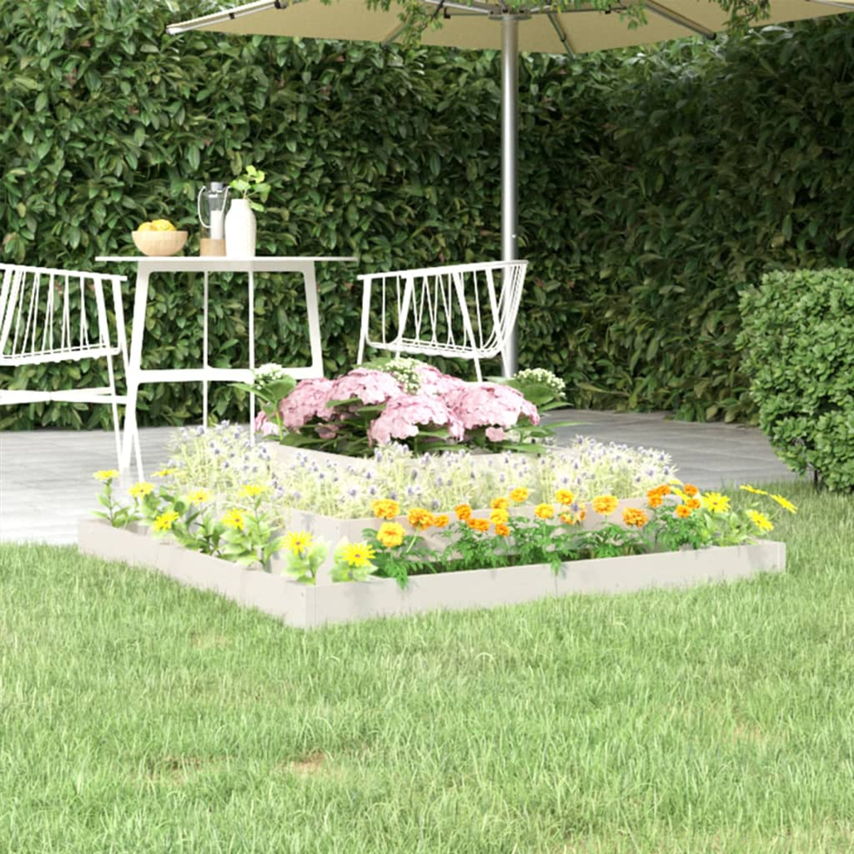 Ein weißes Hochbeet mit Blumen, Tisch, Stühlen und Sonnenschirm auf Gras. Grüne Hecke im Hintergrund.