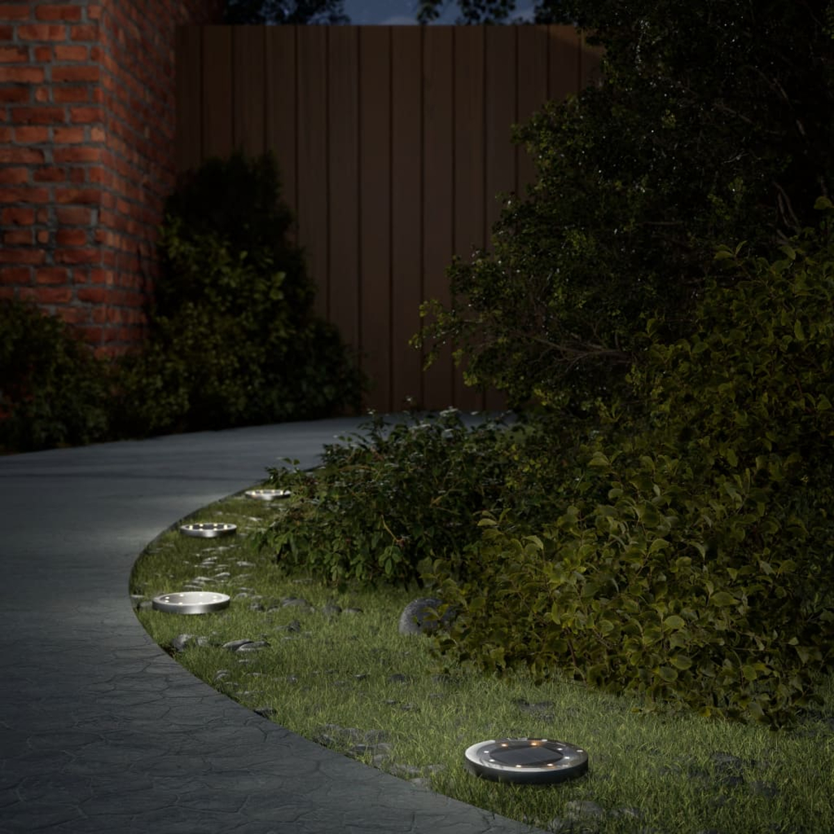 Ein Weg mit Gartenlichtern wird nachts beleuchtet. Büsche und eine Backsteinmauer im Hintergrund.