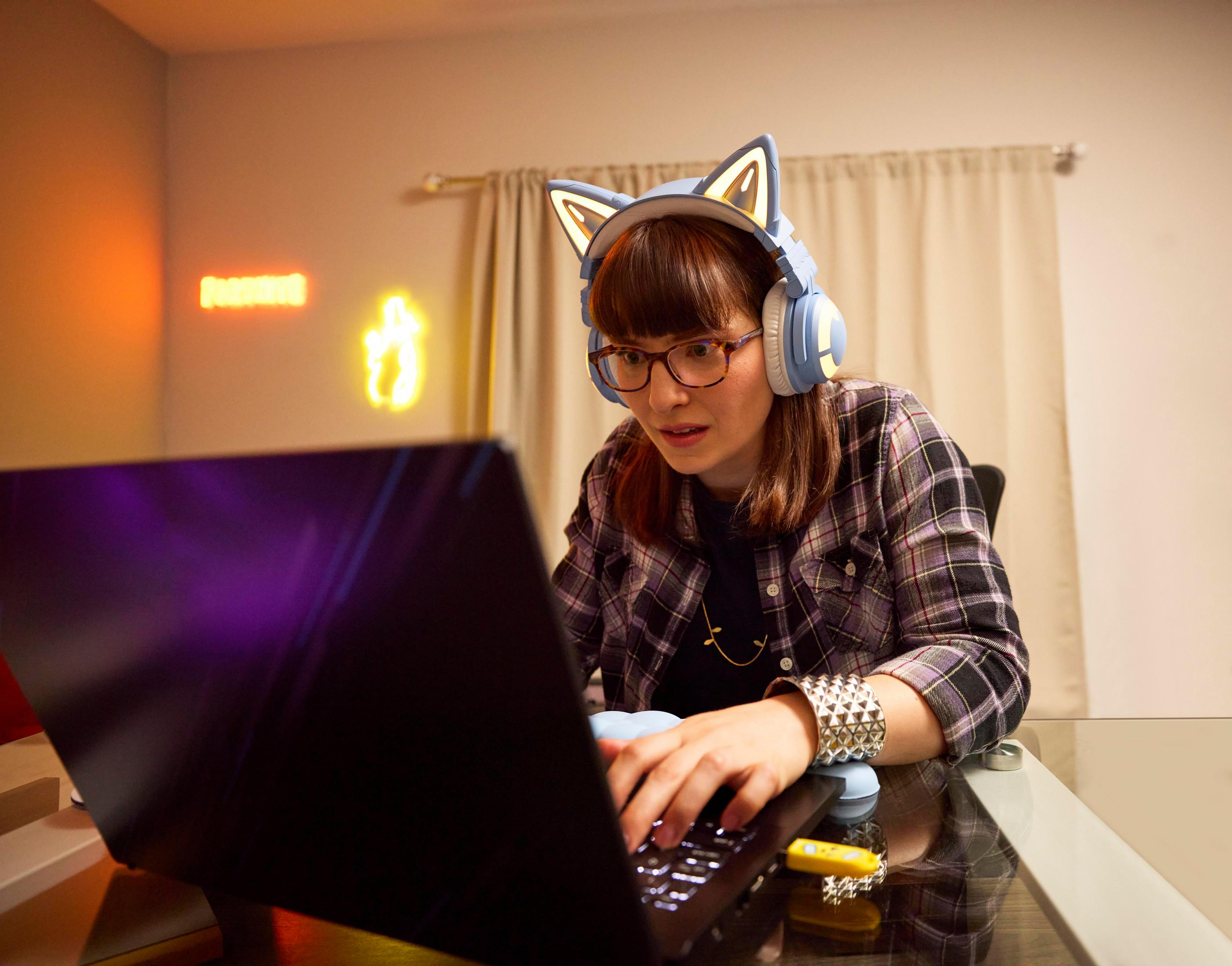 Kobieta ze słuchawkami, patrząca na ekran laptopa, w pokoju z neonami.