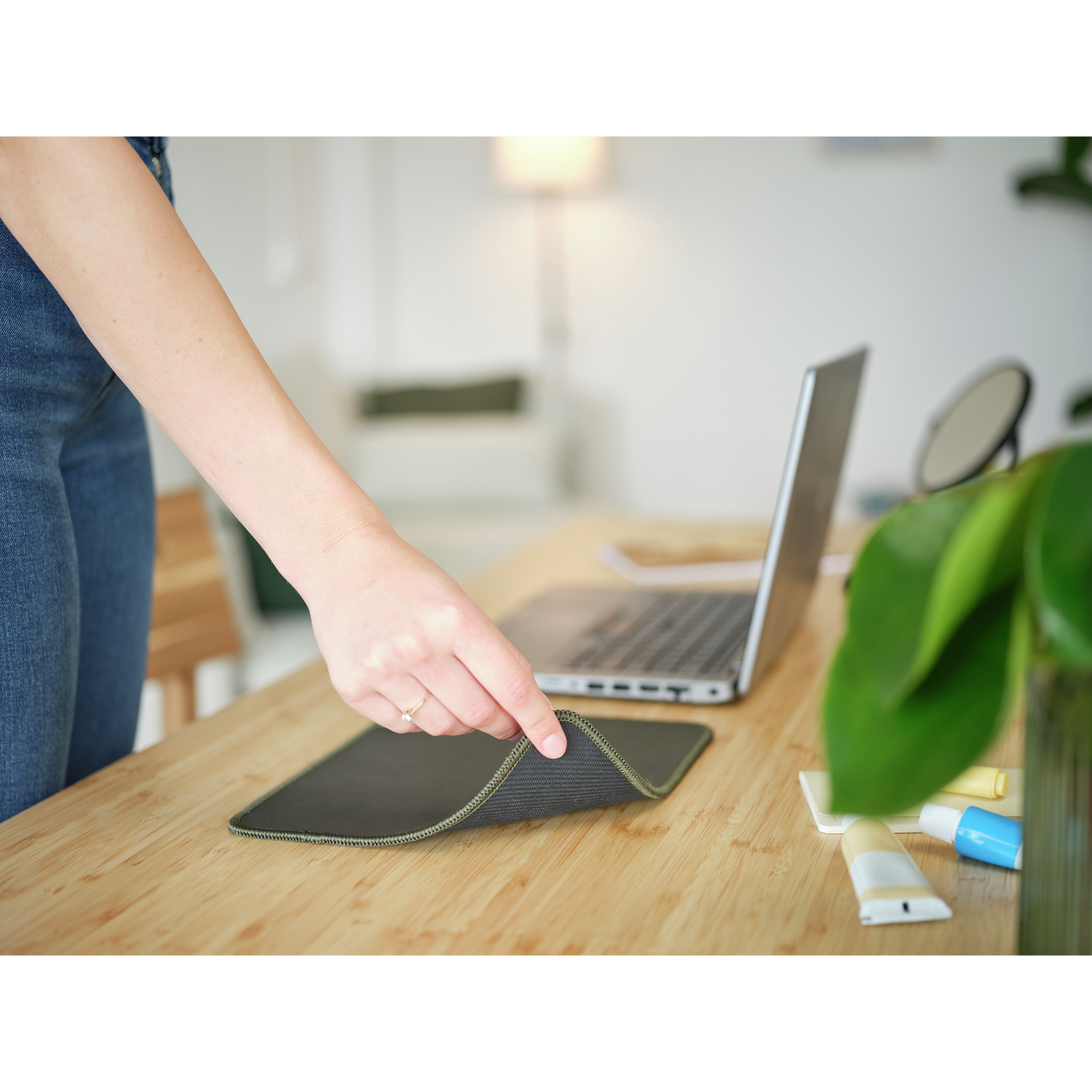 Une main tenant un tapis de souris noir sur un bureau en bois, à côté d'un ordinateur portable et d'une plante.