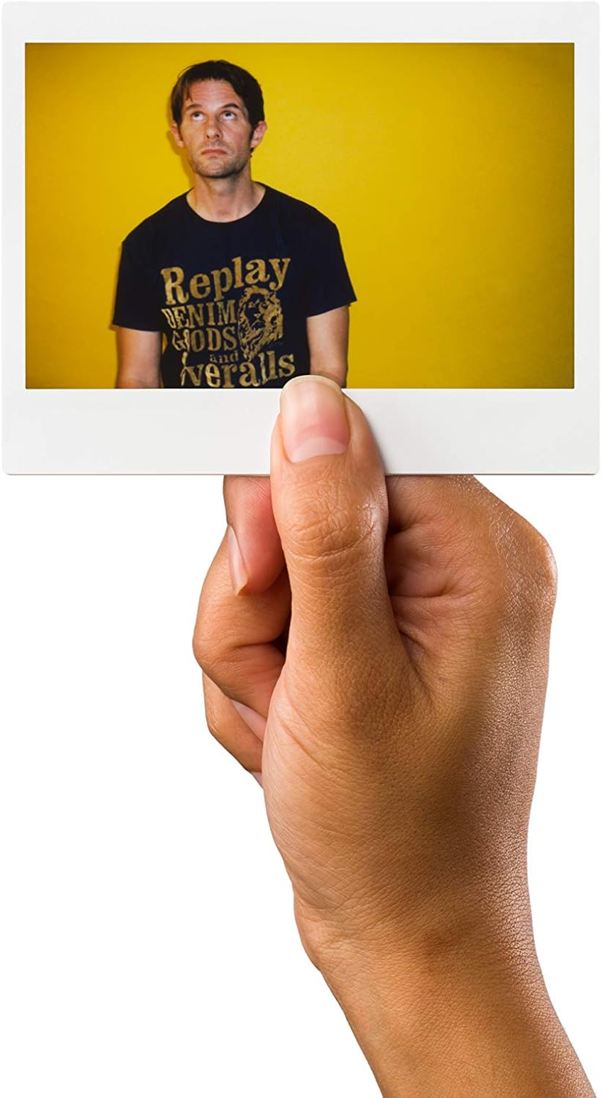 Una mano sosteniendo una foto polaroid de un hombre contra un fondo amarillo. El hombre mira hacia arriba.