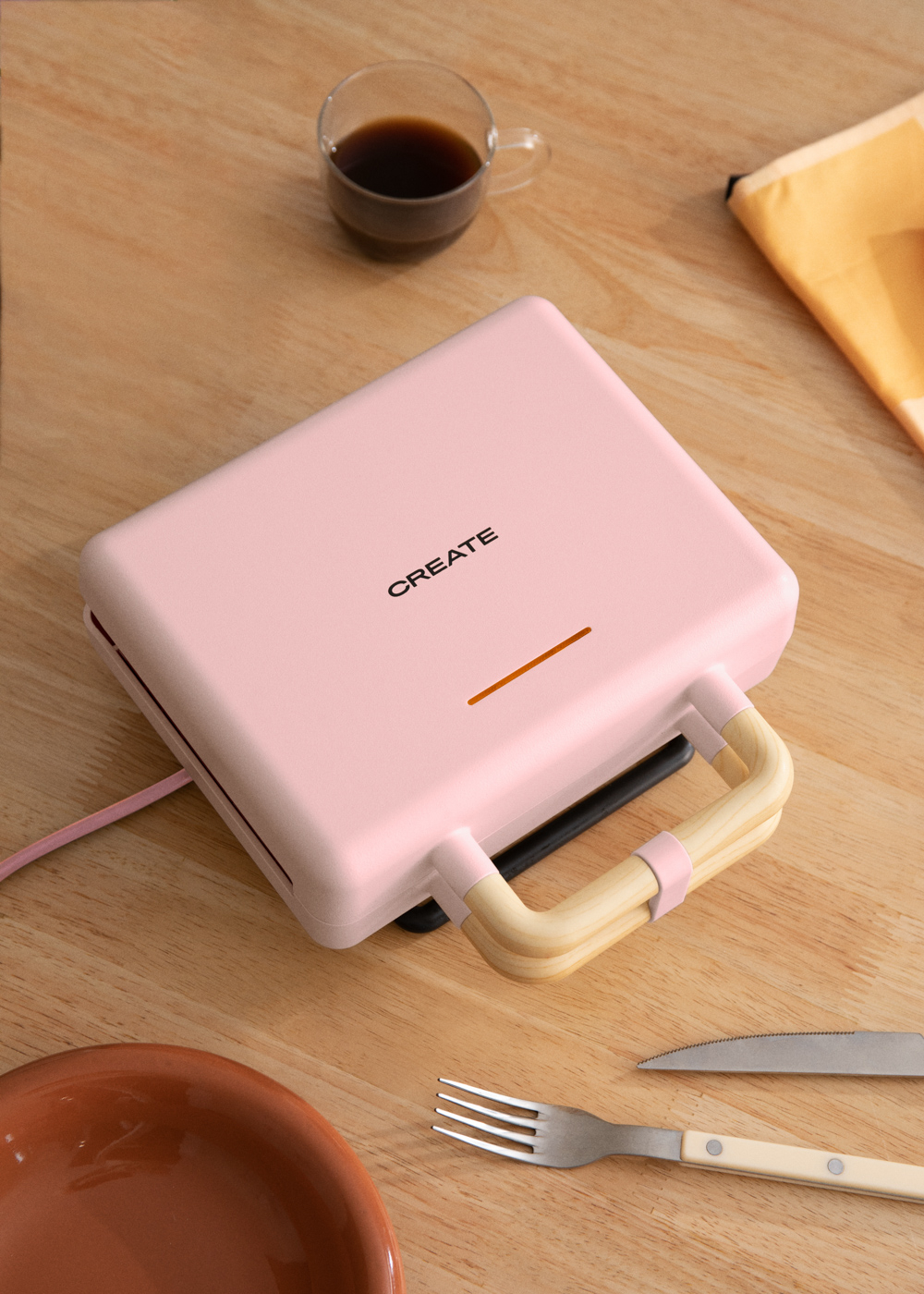 Una sandwichera CREATE rosa sobre una mesa de madera. Una taza de café y utensilios al lado.