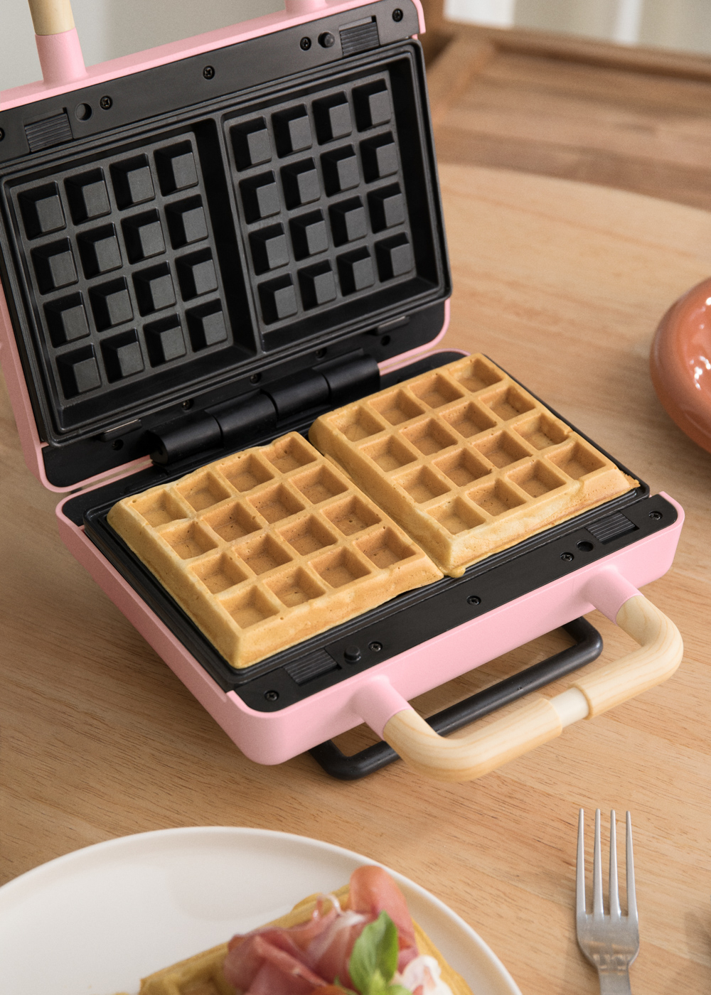 Una gofrera rosa con dos gofres dentro, colocada sobre una superficie de madera.