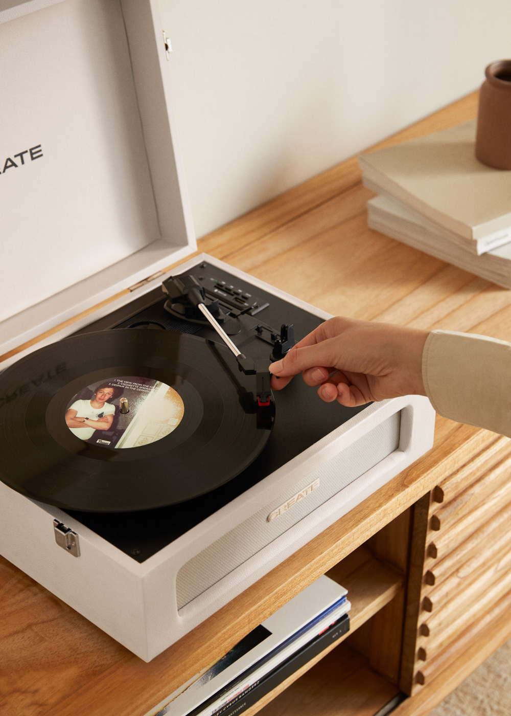 Una mano ajusta un disco en un tocadiscos blanco. CREATE es visible. Mueble debajo.
