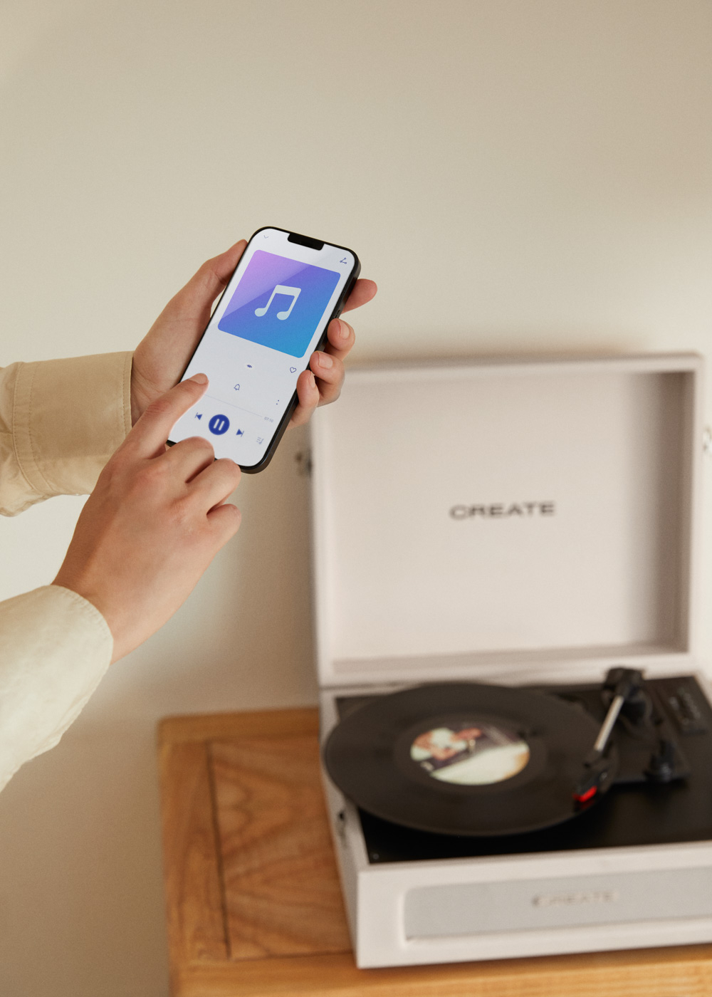 Una mano sostiene un teléfono que muestra controles de música. Hay un tocadiscos blanco cerca. CREATE es visible.