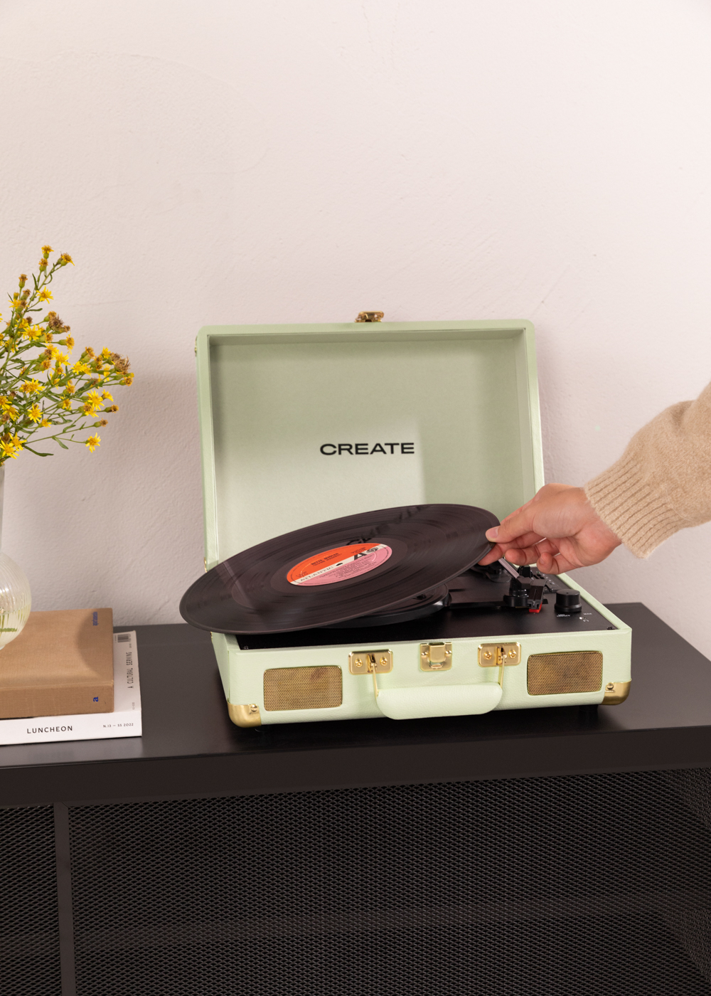 Una mano quita un disco de un tocadiscos verde. CREATE es visible. Mueble negro debajo.