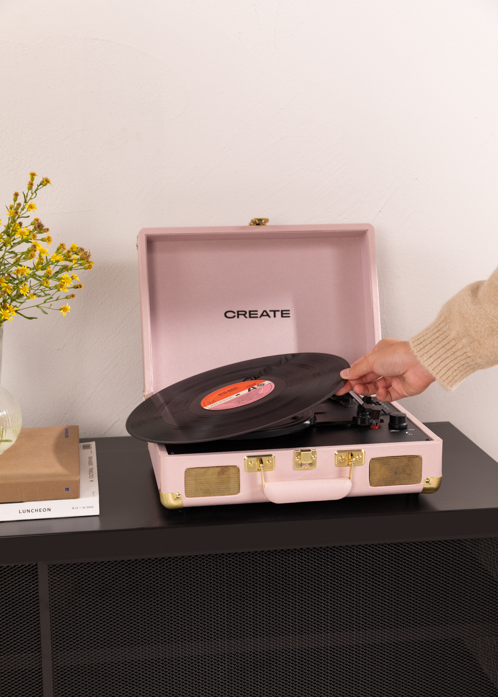 Tocadiscos rosa, una mano pone un disco. Flores y libros sobre la mesa negra.
