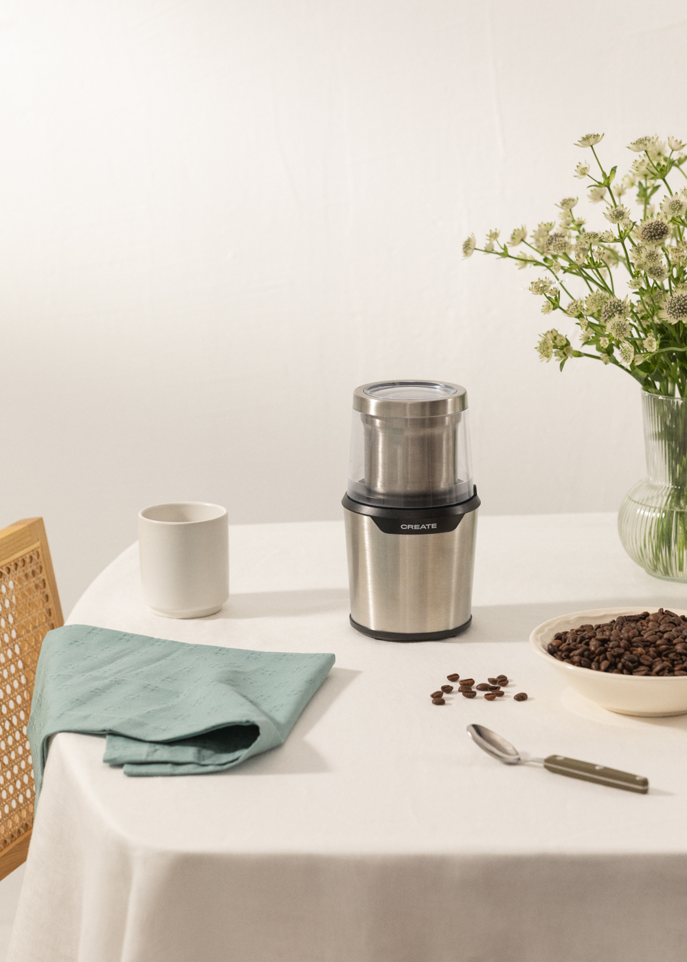 Molinillo de café en una mesa blanca. Granos de café, taza, cuchara y flores visibles.