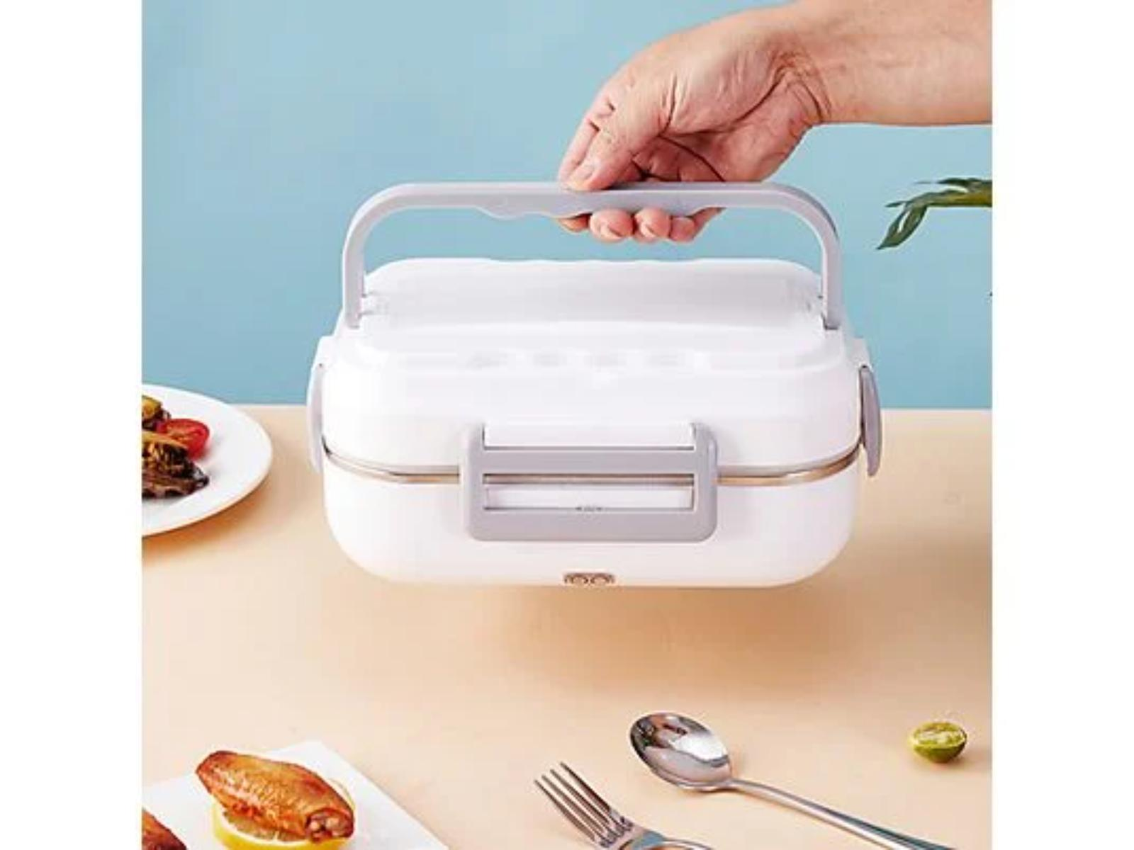 Hand hält eine weiße Lunchbox mit Griff. Ein Teller mit Essen und Besteck befinden sich im Hintergrund.