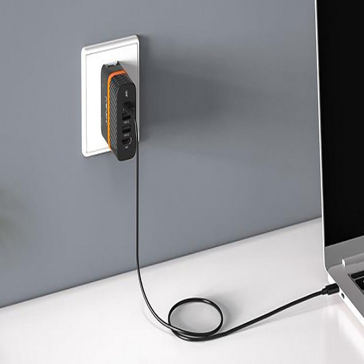 Ein schwarz-oranges Ladegerät in einer Steckdose, mit Laptop und Kabel auf einem weißen Schreibtisch.