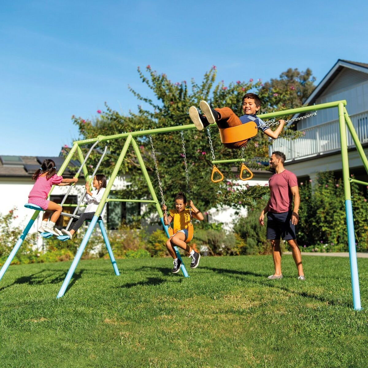 Kinder spielen auf einem grün-blauen Schaukelgerüst. Ein Mann beobachtet. Sonniger Tag, grünes Gras, blauer Himmel.