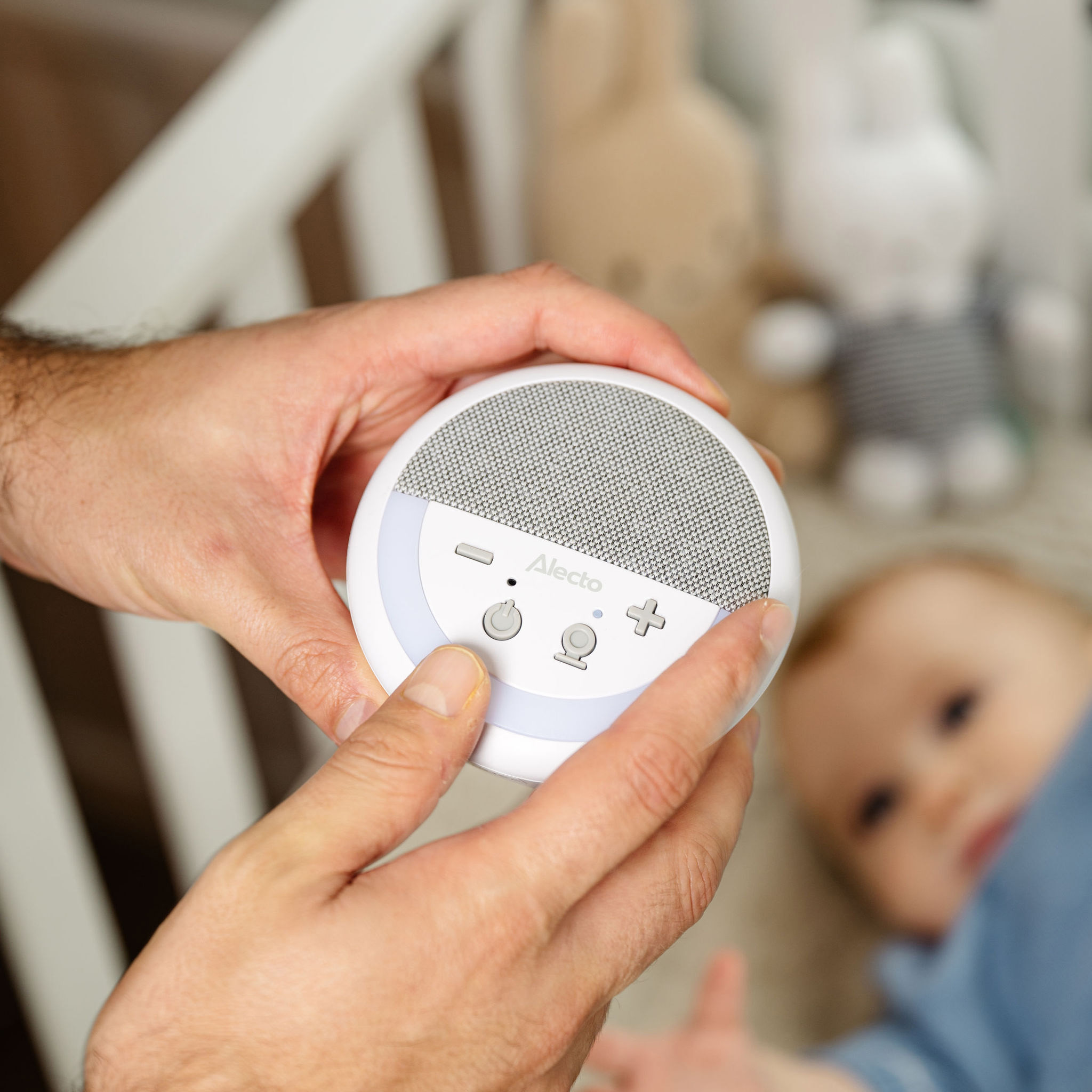 Mains d'une personne tenant une petite machine à sons blanche et grise, un bébé et un berceau en arrière-plan.