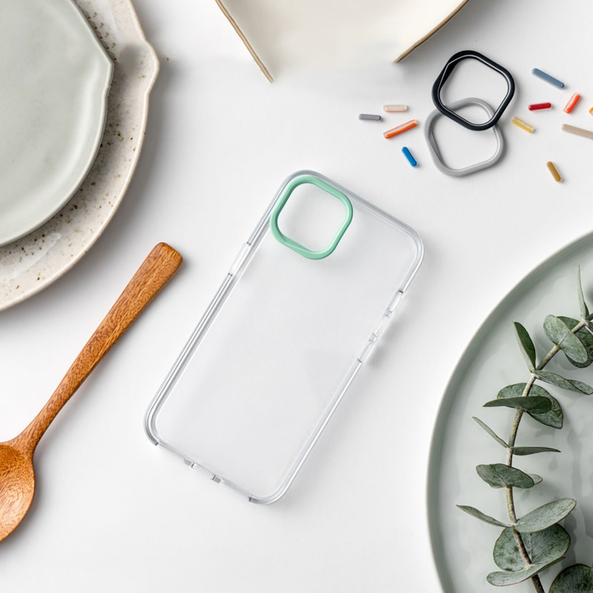 Una custodia trasparente per telefono con un anello per fotocamera verde, vari anelli colorati, un cucchiaio e piatti su una superficie bianca.