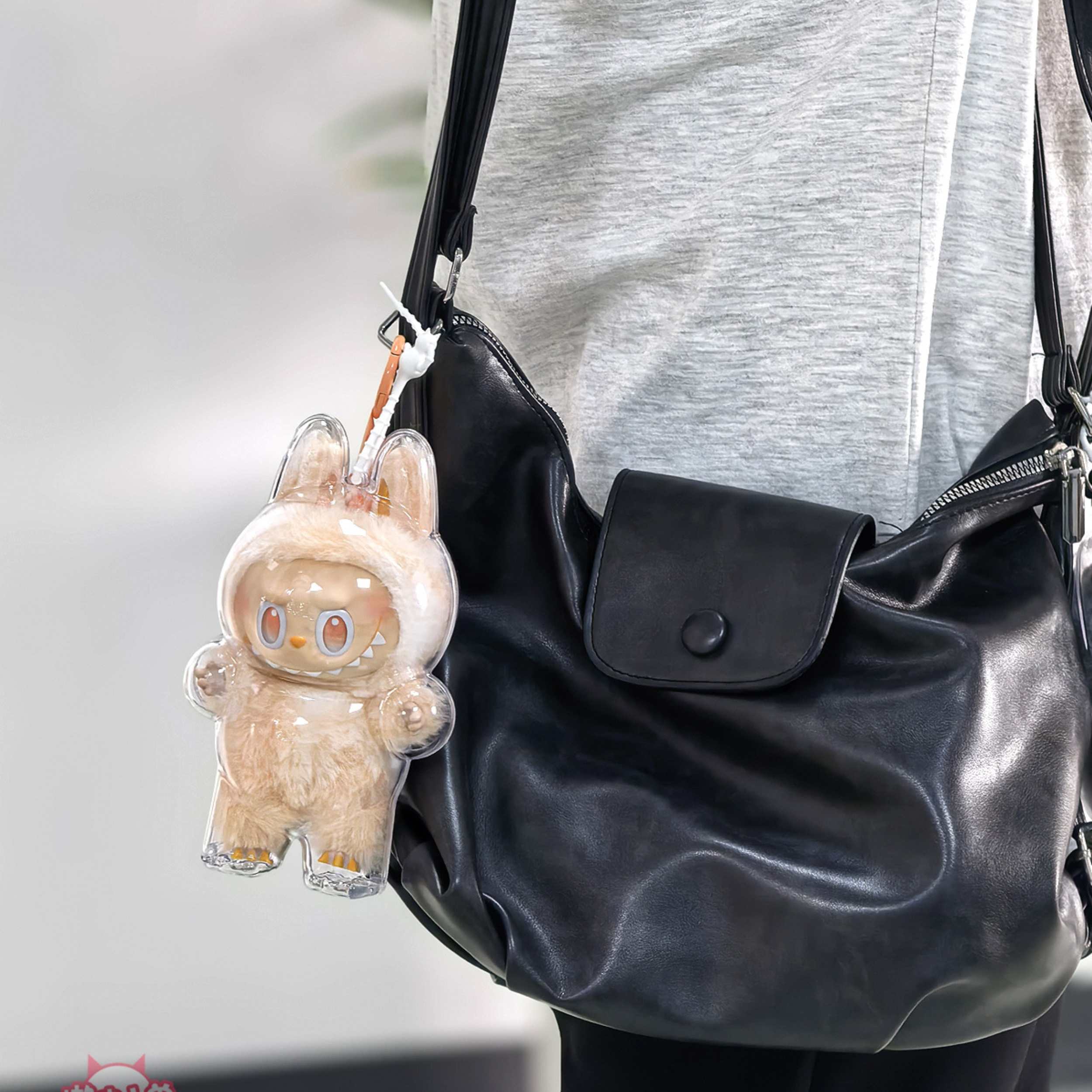 Eine durchsichtige Hasenfigur in einer durchsichtigen Hülle, die an einer schwarzen Umhängetasche hängt, mit grauem Oberteil.