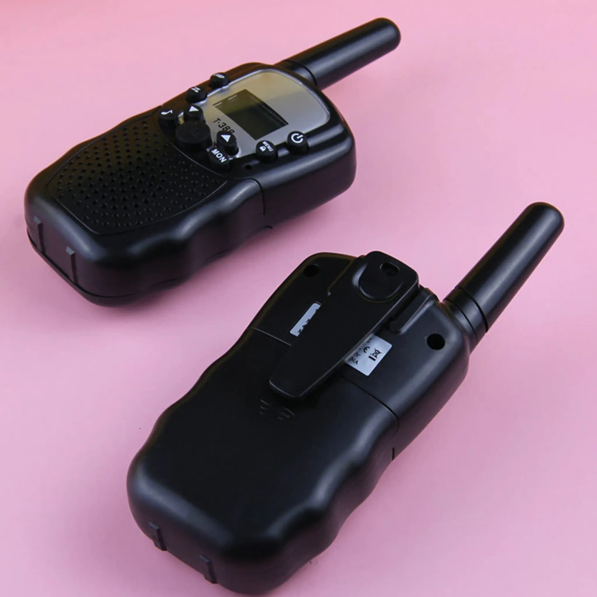 Zwei schwarze Walkie-Talkies auf rosa Oberfläche. Eines zeigt die Rückseite, das andere die Vorderseite.