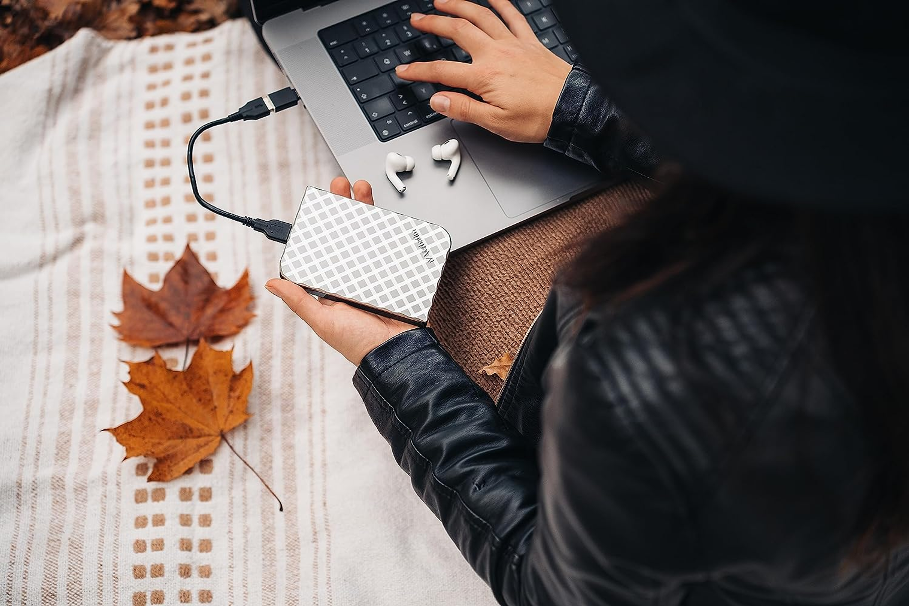 Osoba korzystająca z laptopa, trzymająca zewnętrzny dysk twardy Verbatim. Jesienne liście w tle.