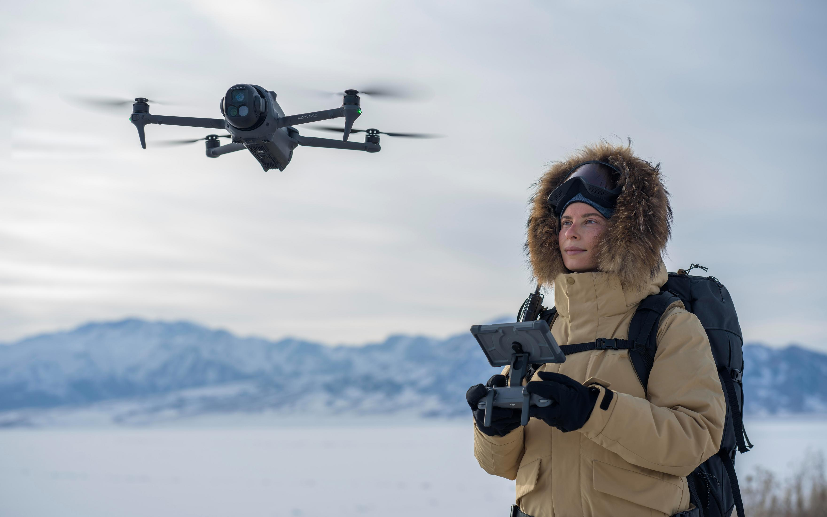 Kobieta obsługująca drona w zaśnieżonym krajobrazie. Dron w locie. Góry w tle.