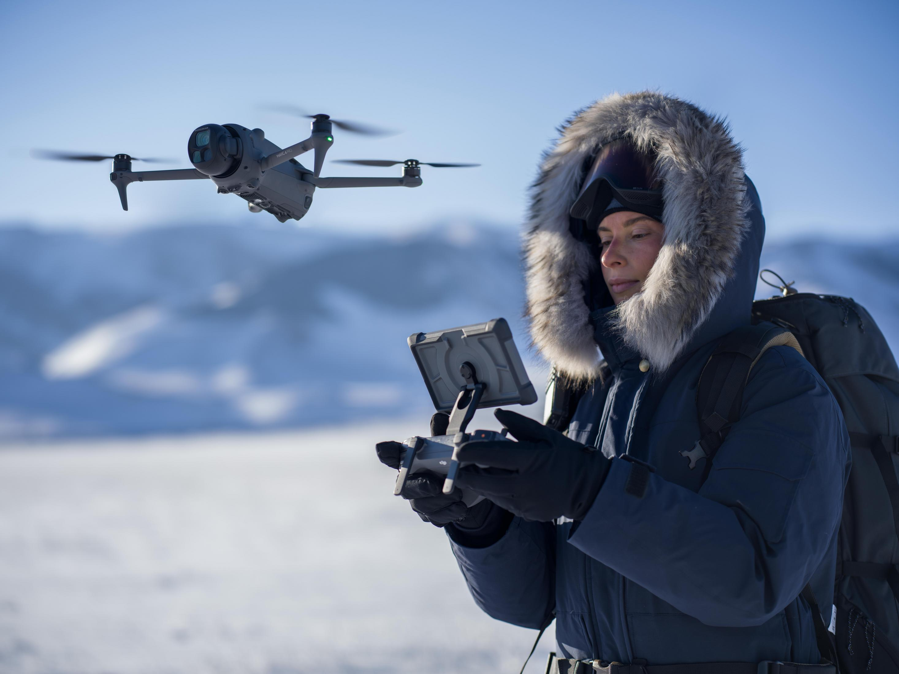 Kobieta w kurtce kontroluje drona na śnieżnym krajobrazie; dron w powietrzu.
