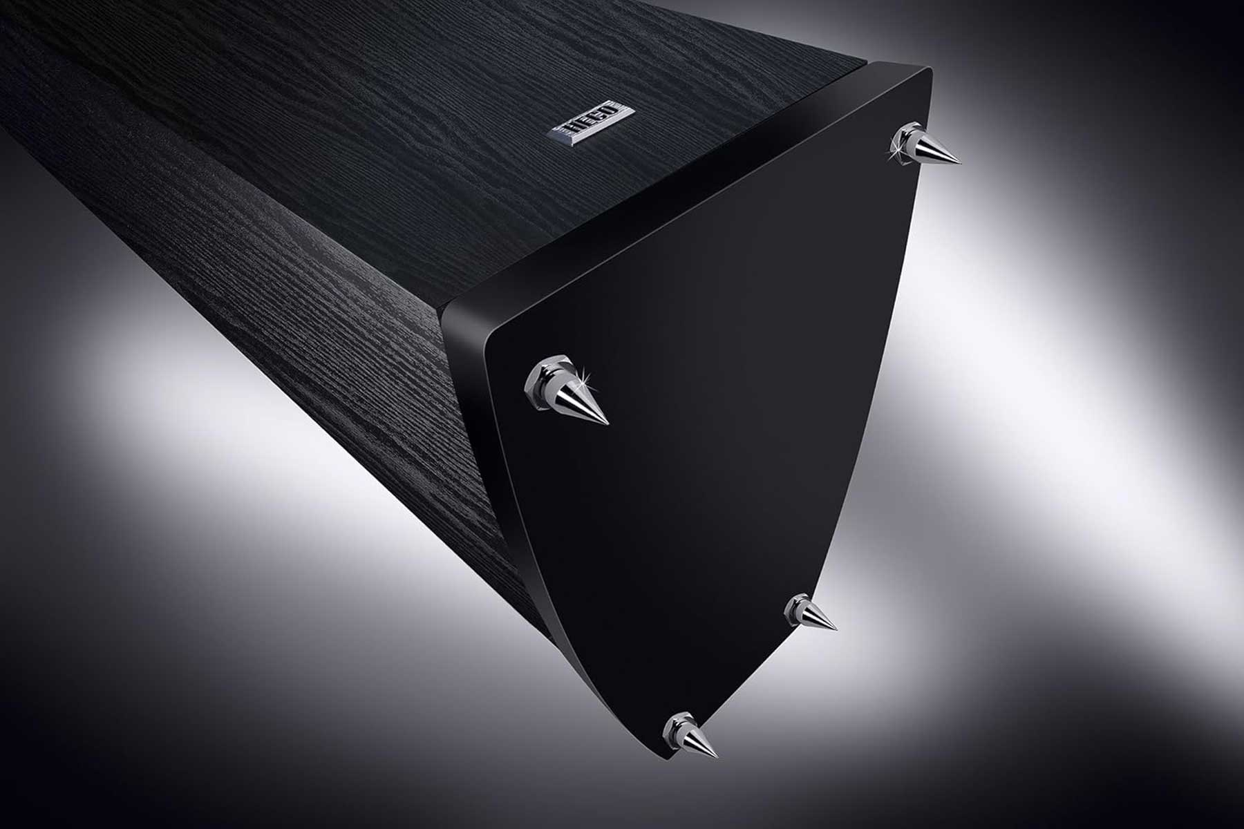 Schwarzer Lautsprecher mit silbernen Spikes auf grauem Hintergrund. Scharfe Winkel und schlankes Design.