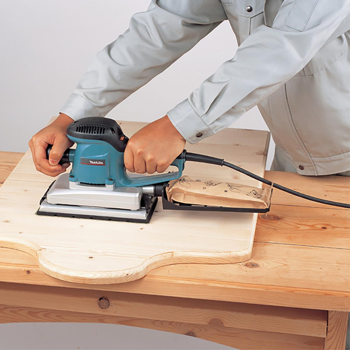 Una persona lija una tabla de madera con una lijadora Makita. La lijadora es azul y negra, la tabla marrón claro. Mesa de madera.