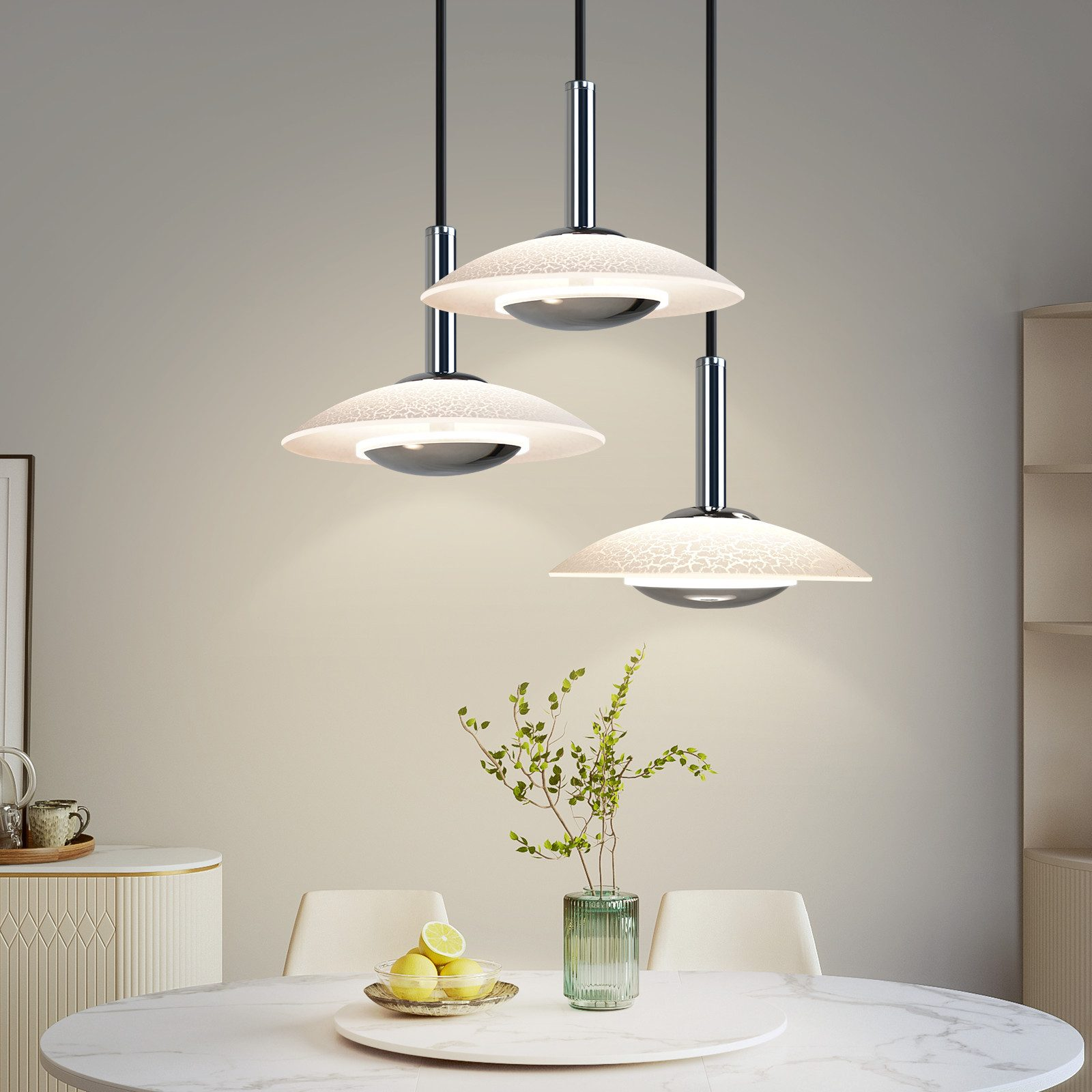 Drei Pendelleuchten mit Milchglasschirmen hängen über einem Esstisch mit einer Blumenvase und Zitronen.
